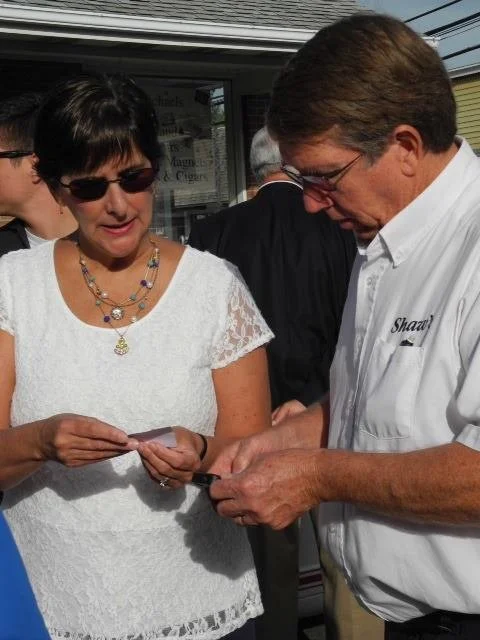 A woman wearing sunglasses and a white lace top looks at a piece of paper while a man in a white shirt with embroidered text and glasses shows her something.