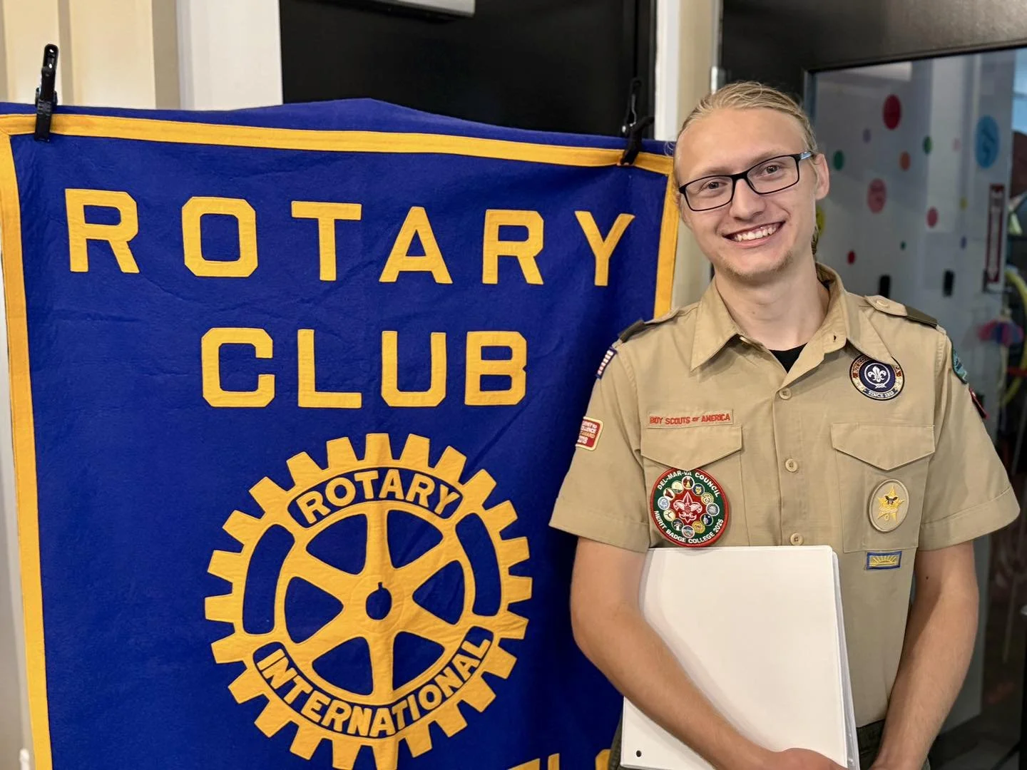 A young man in Boy Scout uniform standing next to a blue Rotary Club banner with yellow text and Rotary gear logo. He is holding a white folder and smiling.