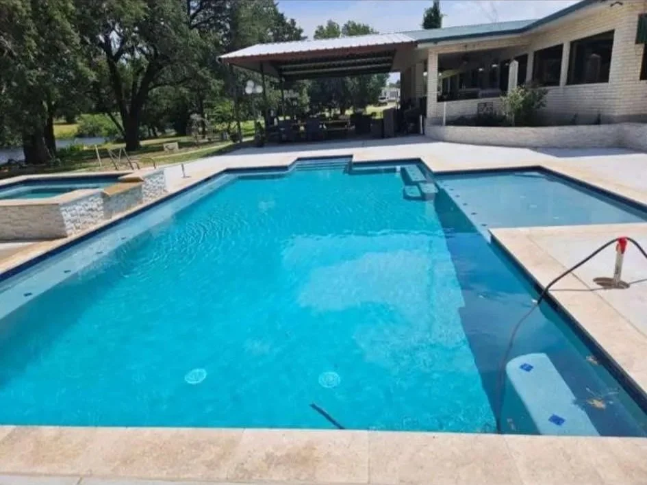A swimming pool with a connected hot tub in a backyard setting, surrounded by a concrete deck, with trees and a house in the background.
