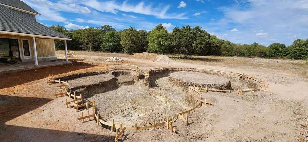Under construction building site with large oval-shaped foundation outline in dirt and gravel, lined with wooden stakes and planks, next to a yellow house with a porch and trees in the background, under a partly cloudy sky.