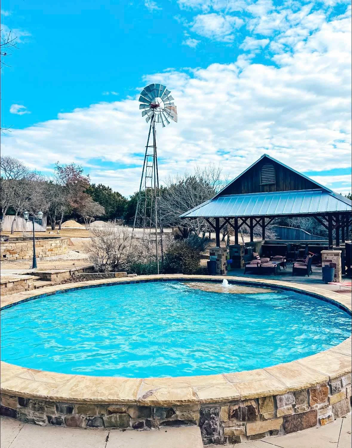 A backyard with a circular swimming pool, a windmill, a covered patio with outdoor furniture, and trees under a partly cloudy blue sky.