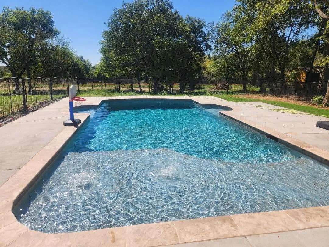 A backyard swimming pool with a shallow area, surrounded by a concrete deck and a black metal fence, with trees and a clear blue sky in the background.