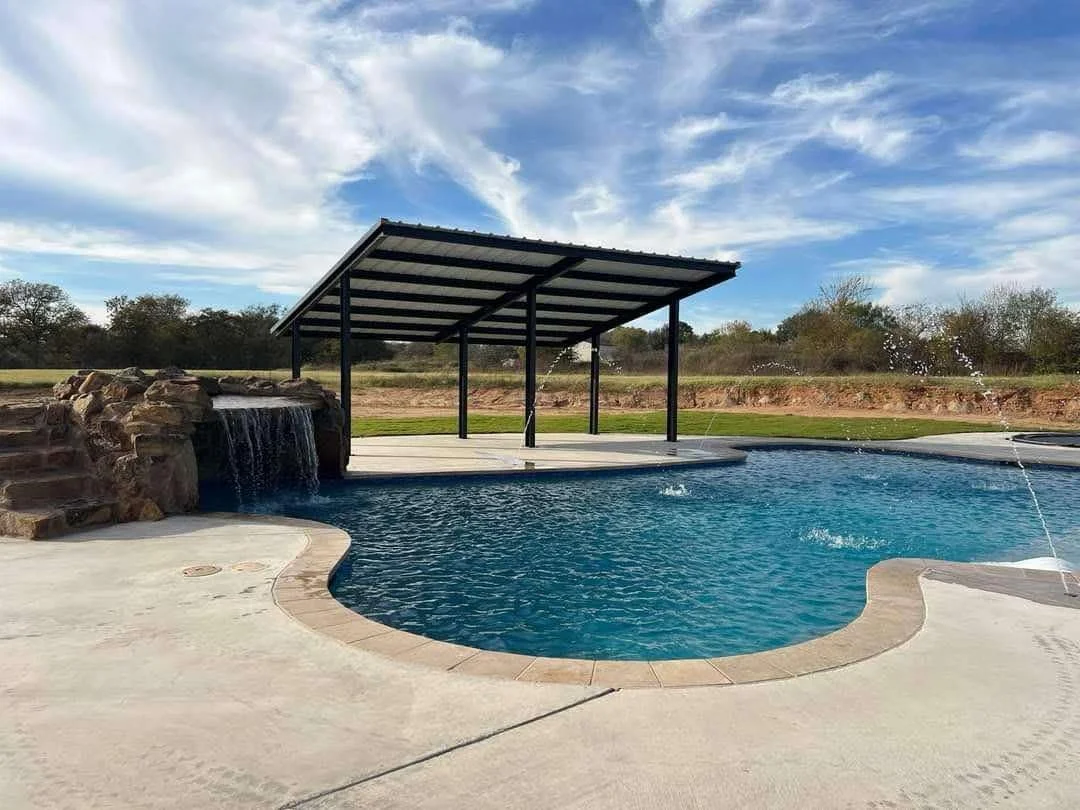 A backyard swimming pool with a waterfall feature, covered patio, and open sky.