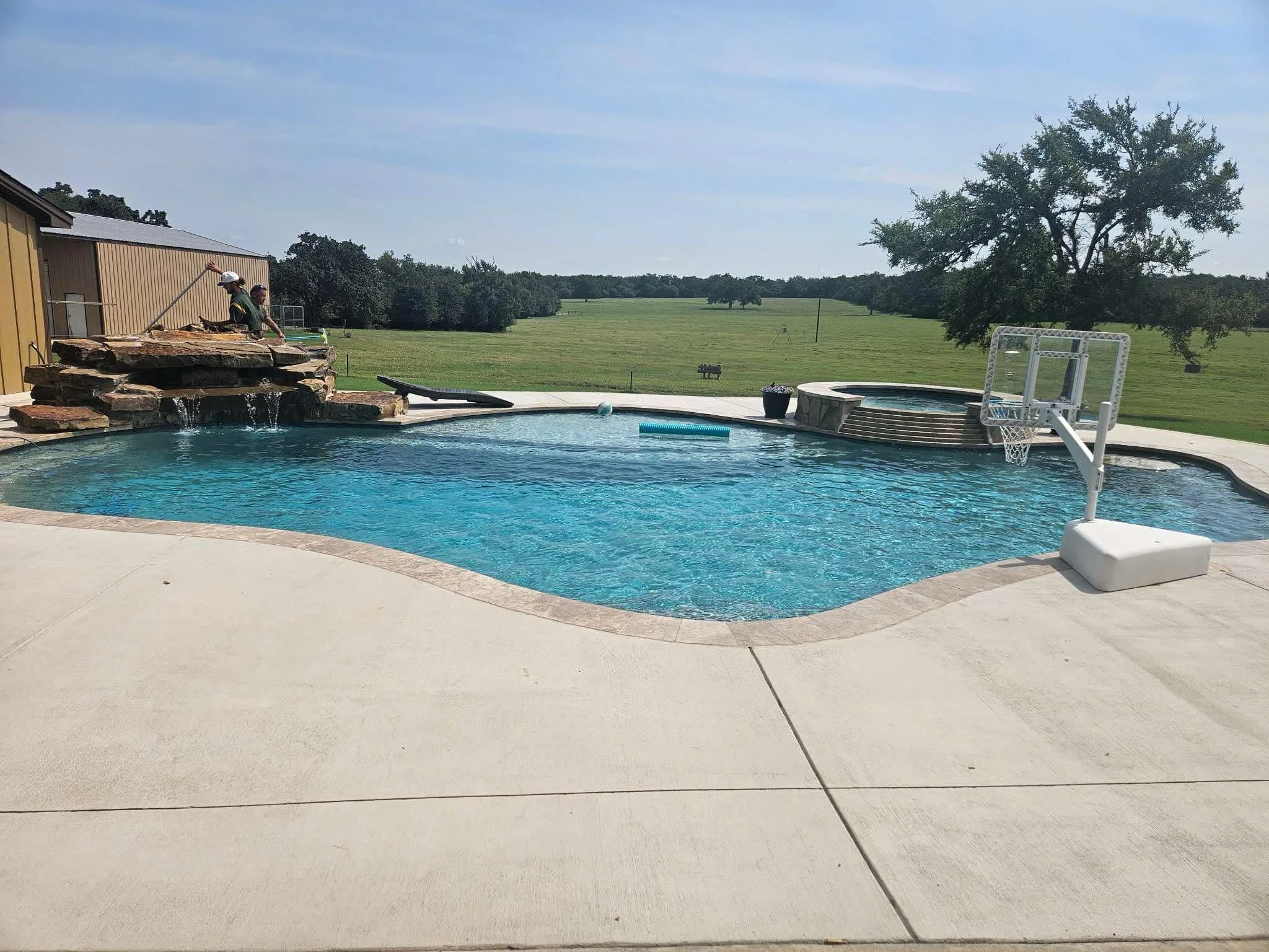 A backyard swimming pool with a rock waterfall feature, a basketball hoop in the water, and a hot tub in the background. Two people are working on the waterfall feature on the left side. The area is surrounded by green open land and a few trees, unde