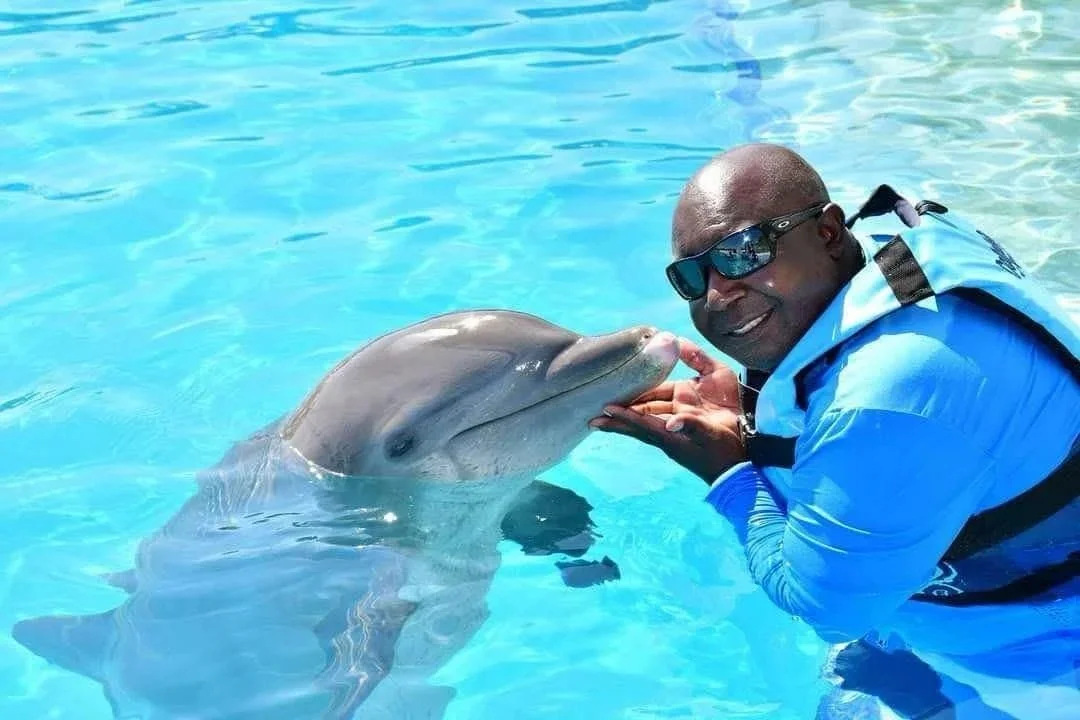 A man wearing sunglasses and a blue life jacket smiling while holding a dolphin close to his face in a bright blue pool.