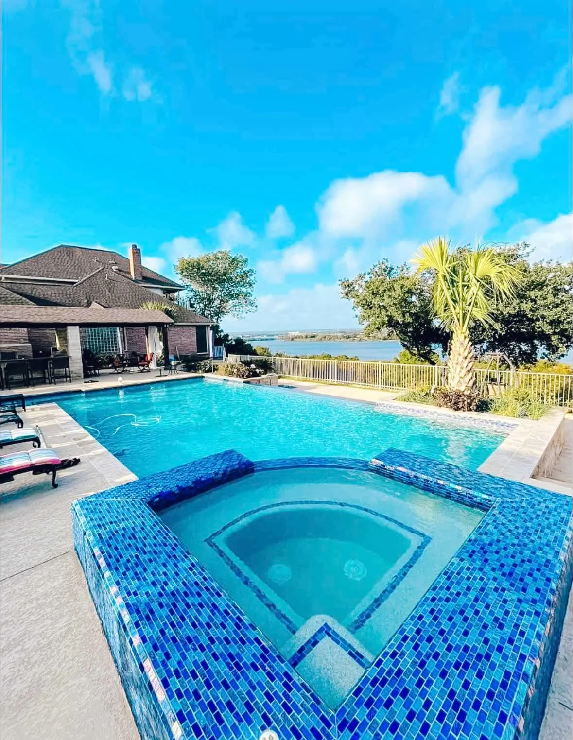 Peaceful backyard with a swimming pool and hot tub overlooking a body of water under a blue sky with scattered clouds.