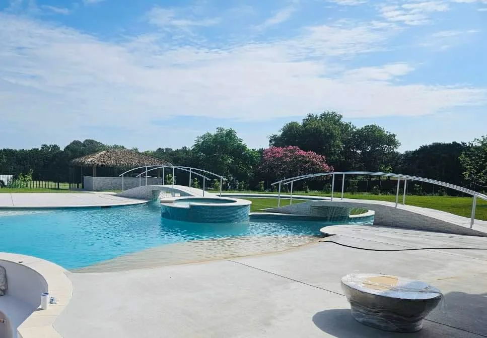 An outdoor swimming pool with a small round hot tub on a bright, sunny day, surrounded by green grass and trees, with a thatched-roof gazebo in the background.