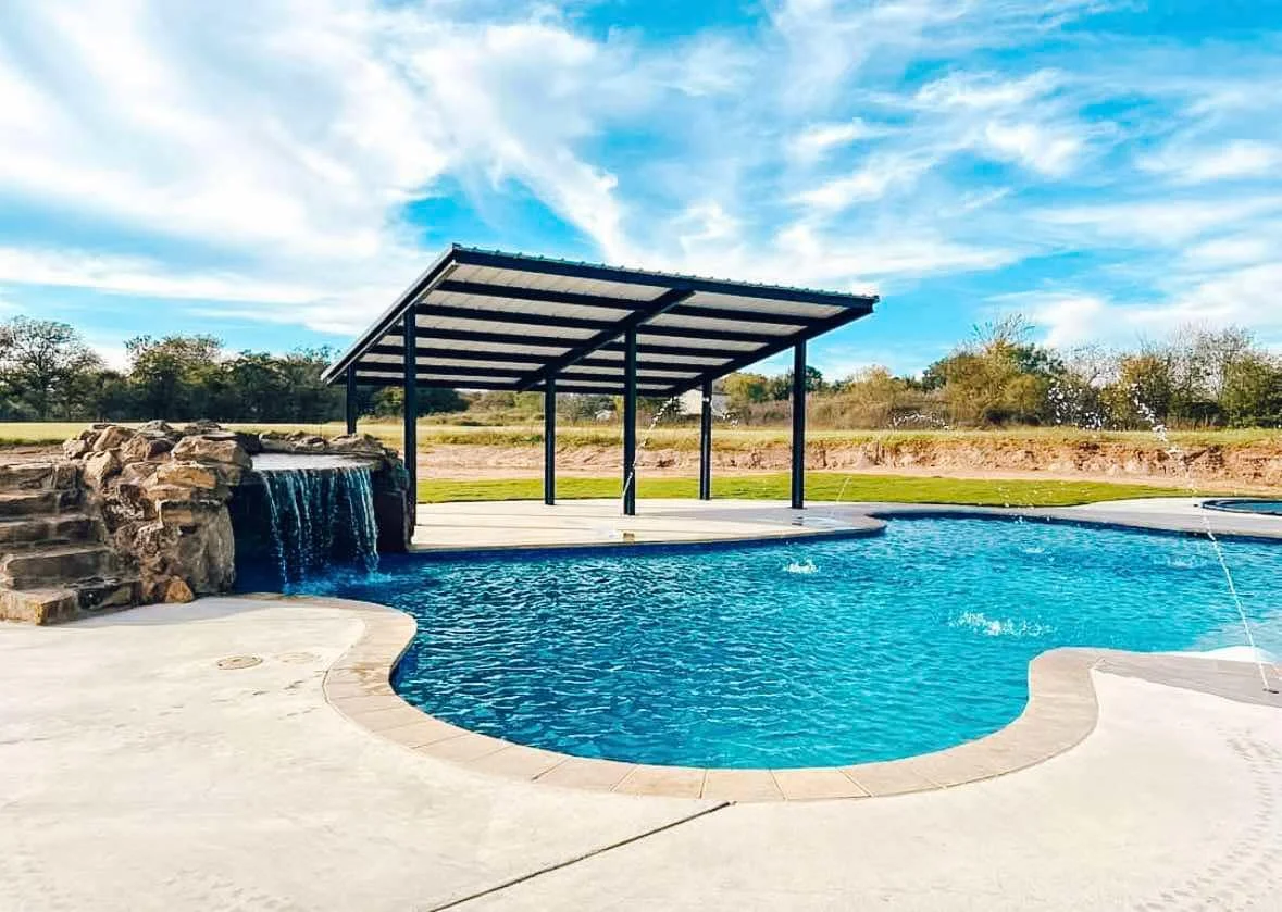A backyard swimming pool with a small waterfall, concrete decking, a shaded pavilion, and a grassy landscape with trees in the background under a partly cloudy sky.