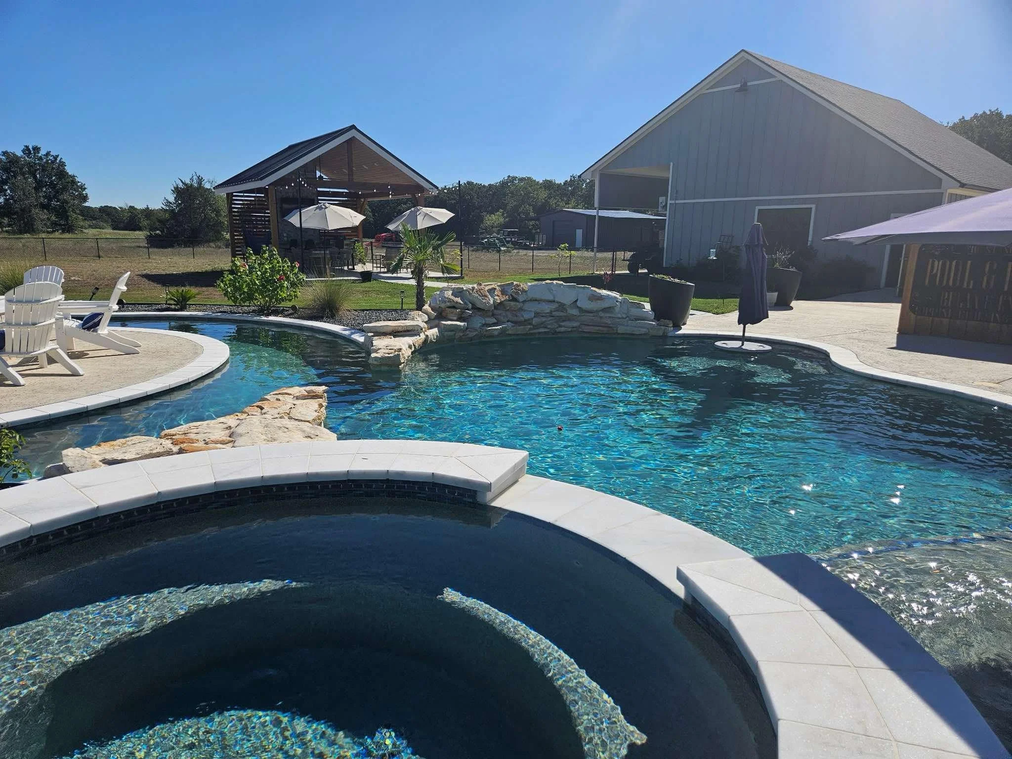 Outdoor pool with a hot tub in the foreground, surrounded by white patio chairs and umbrellas, with a grassy area and a large barn-style building in the background.