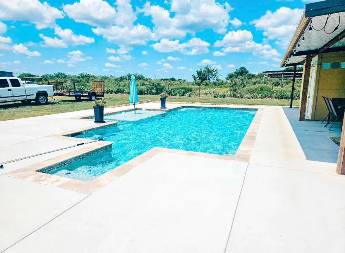 Backyard swimming pool with a clear blue sky and white clouds, surrounded by a concrete deck, a closed blue umbrella, and some potted plants. A white truck and trailer are parked on the lawn, with trees and open land in the background, and part of a 