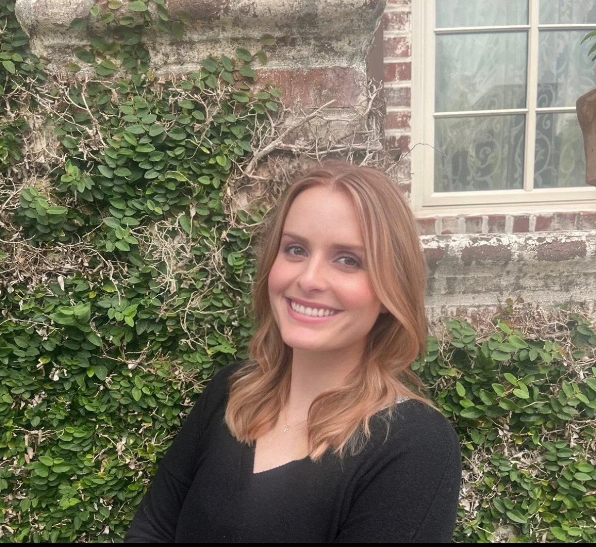 A smiling woman with shoulder-length light brown hair, wearing a black top, standing outdoors in front of a brick wall with ivy and a window.