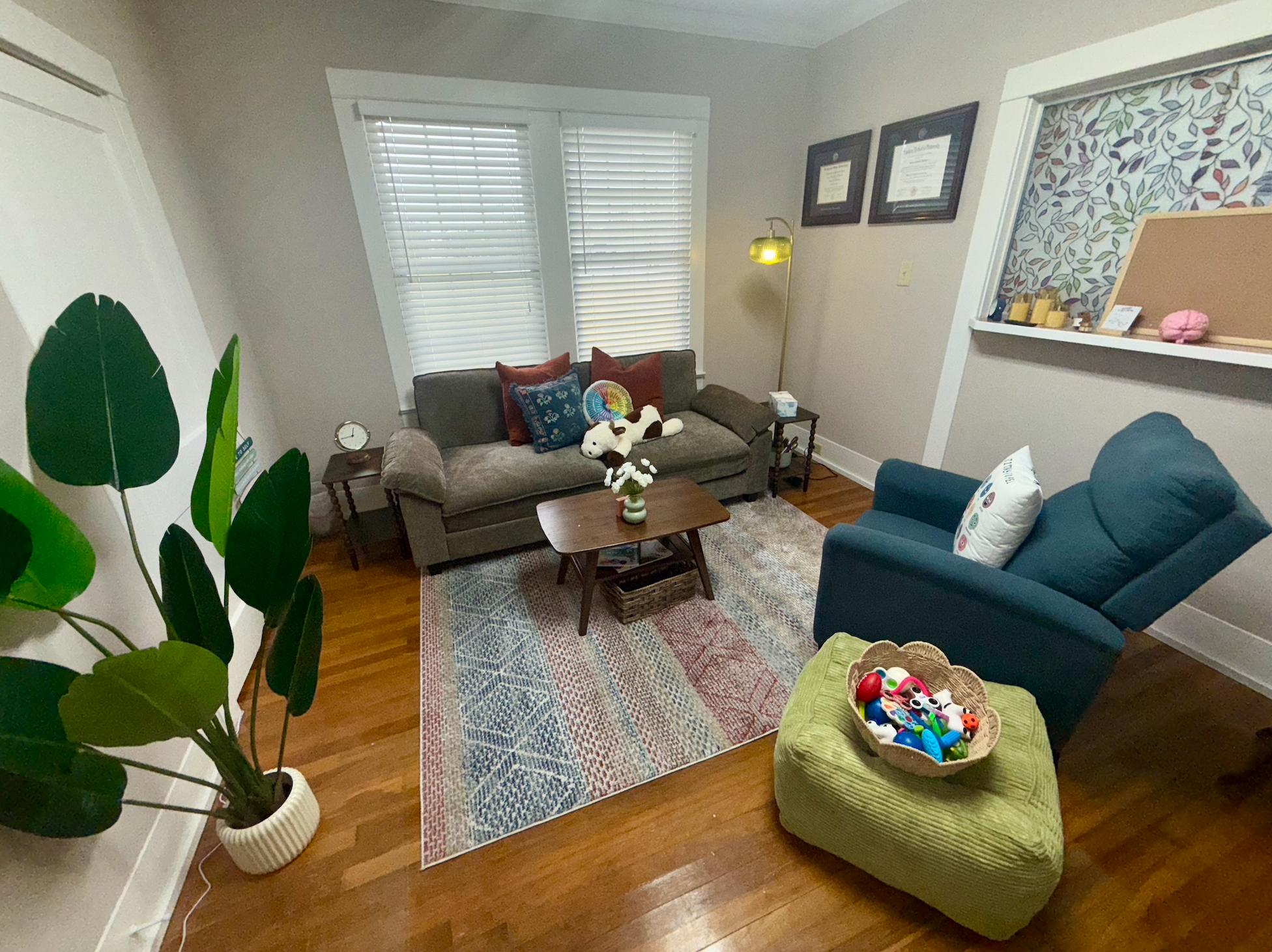 Living room with gray sofa, blue armchair, green ottoman filled with children's toys, wooden coffee table, patterned area rug, houseplant, windows with blinds, framed certificates, floor lamp, shelves with decorative items, and cozy decorative pillows and stuffed animals.