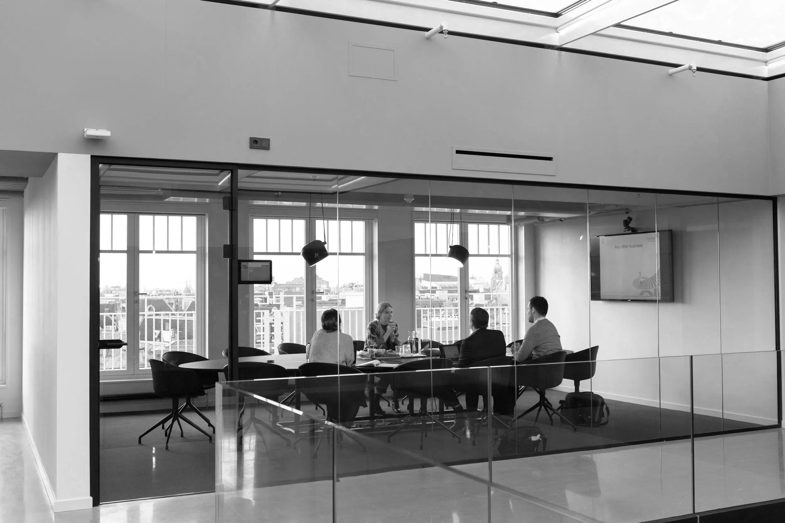Four people seated at a conference table in a bright office meeting room with glass walls, overlooking a city skyline, with a television screen displaying a presentation.