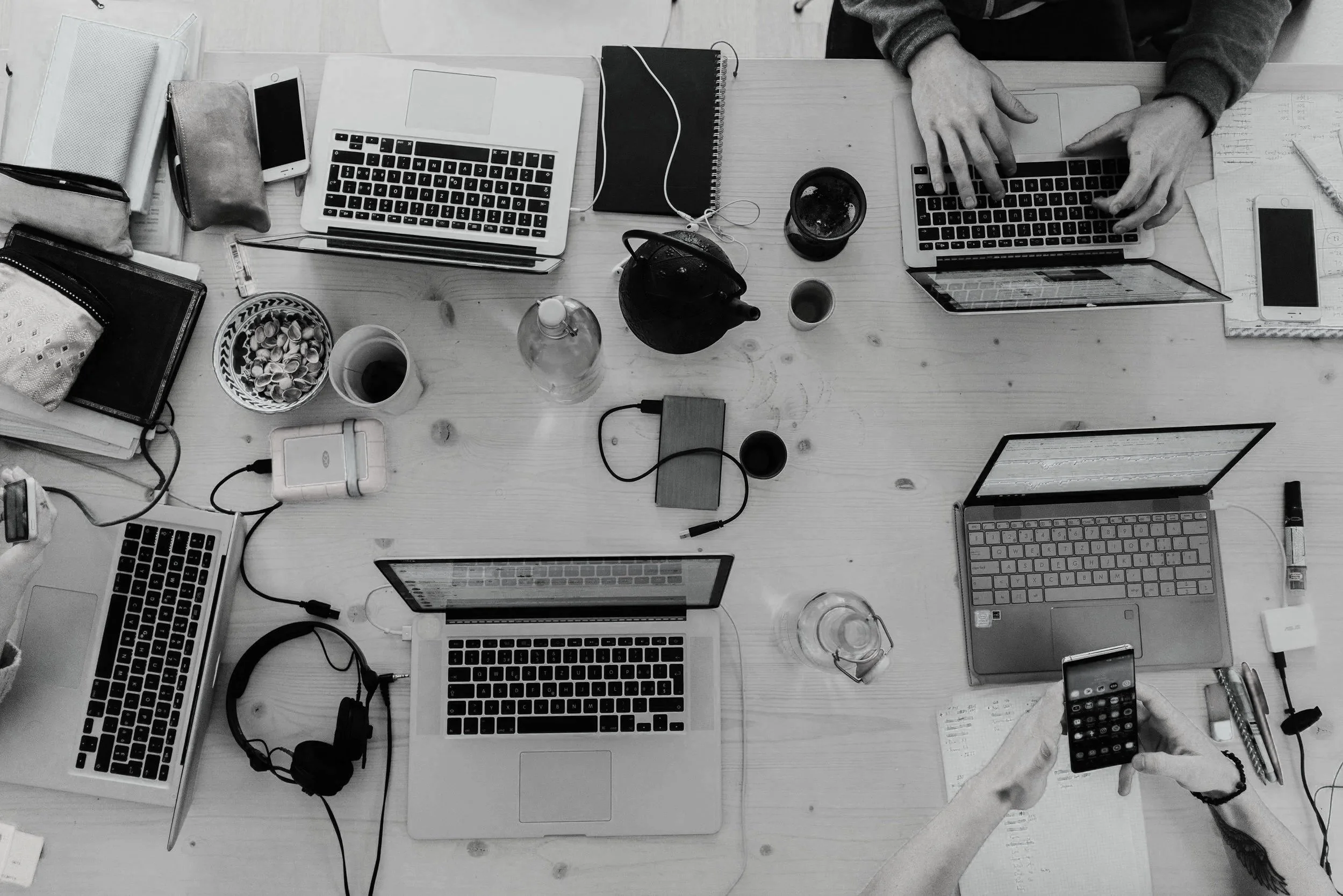 Top-down view of a cluttered wooden table with three laptops, smartphones, notebooks, cups, headphones, a teapot, and various office supplies and personal items. One person is using a laptop on the right side of the image, and another person is holding a smartphone at the bottom.