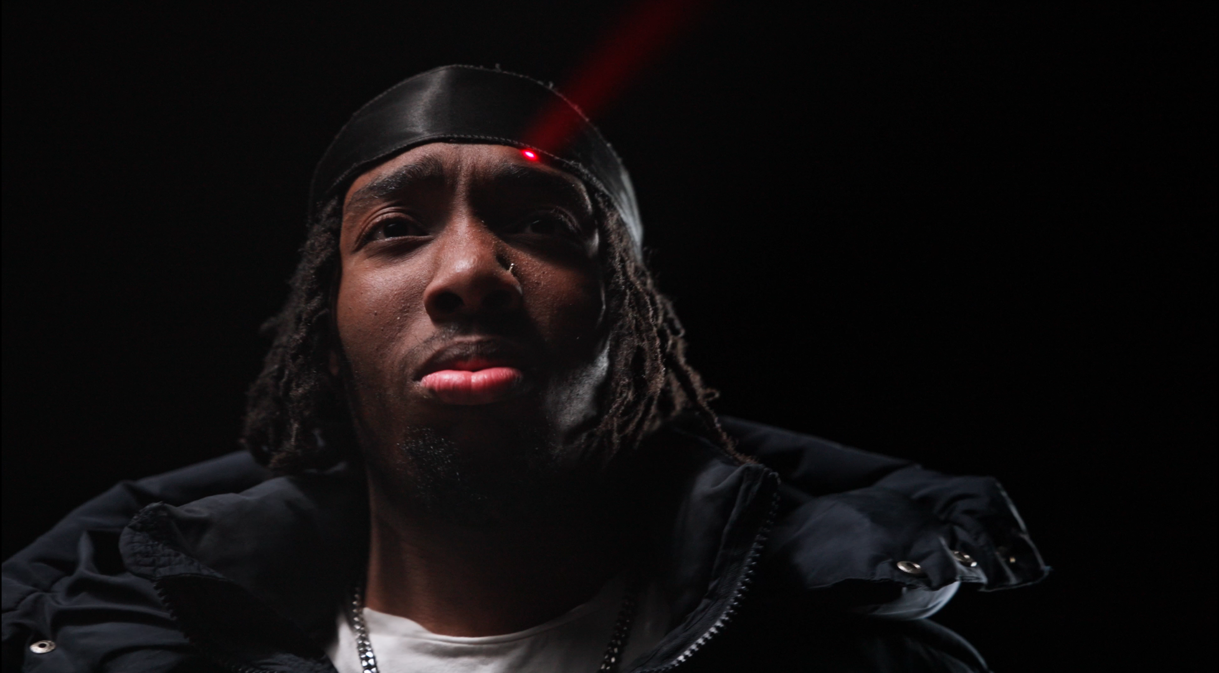 Close-up portrait of a young man with dreadlocks, wearing a black headband, black jacket, and chain, against a dark background.