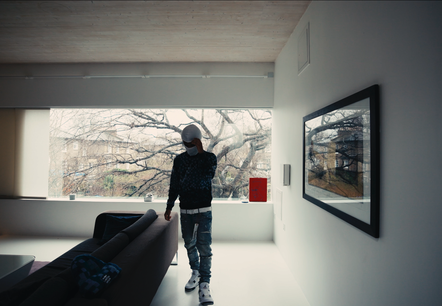 Person in a black patterned sweater, distressed jeans, and white sneakers stands inside a modern living room, adjusting a white face mask and helmet near a large window with leafless trees outside.