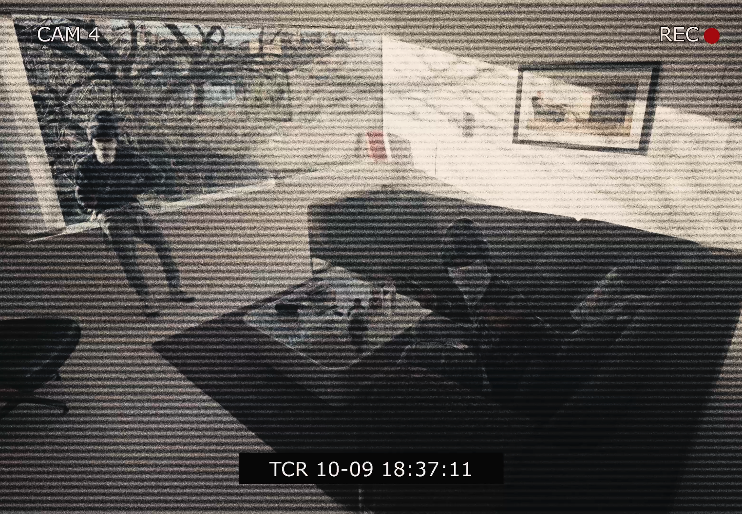 A black-and-white security camera footage showing a person standing in a living room near a door with a painting hanging on the wall, and another person sitting at a desk or table.