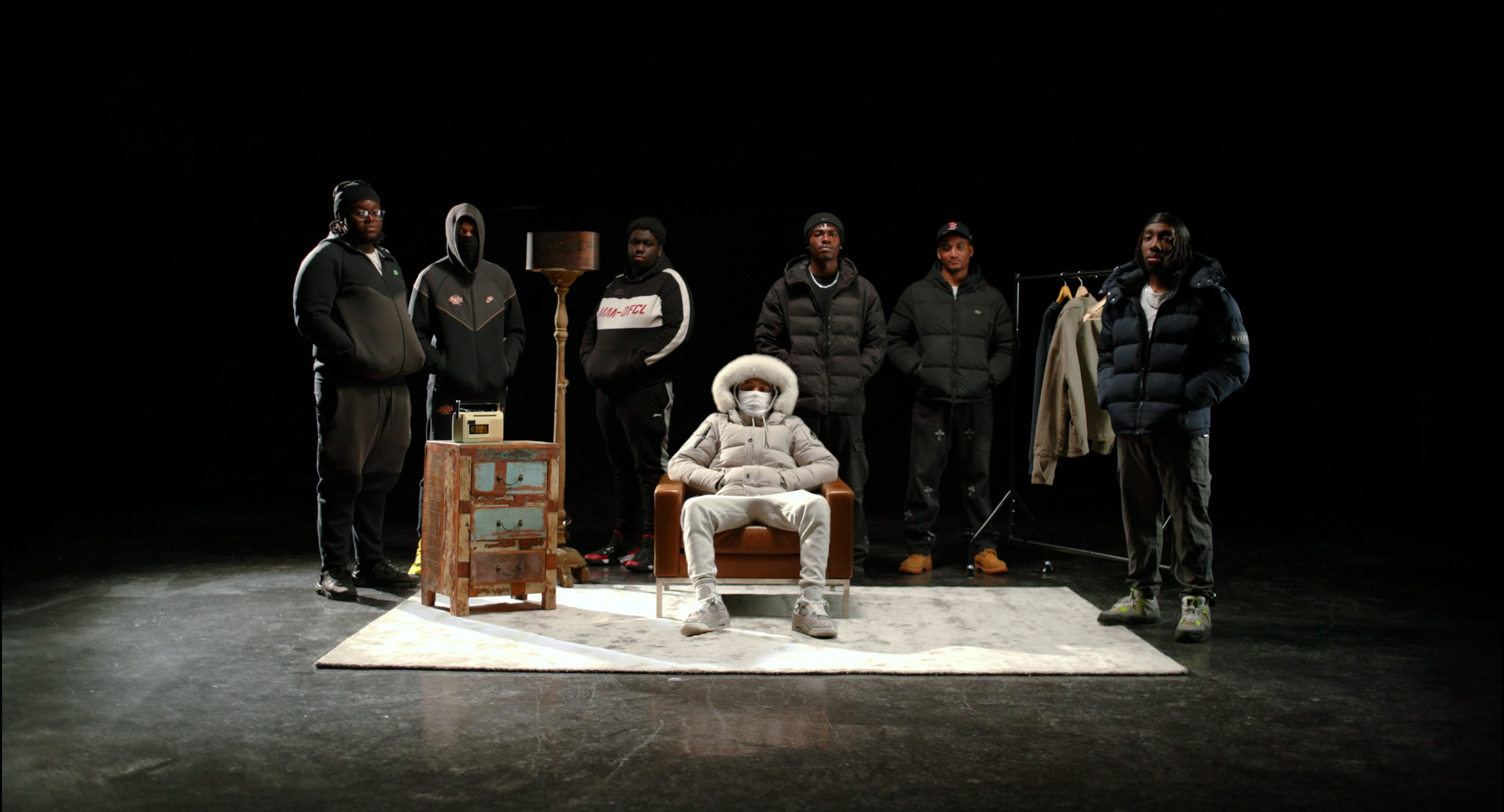 Group of seven young people, six males and one female, in a dark studio setting. The female is seated in a chair in the center wearing a white winter coat with a fur-lined hood and a face mask. The males are standing around her, dressed in casual win