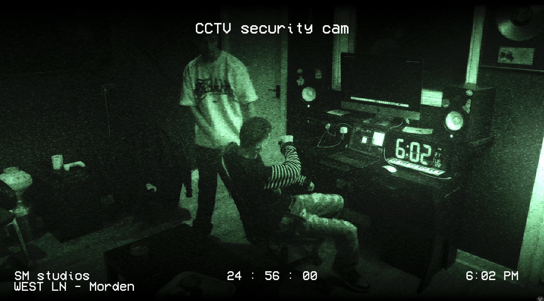 Night vision security camera footage showing two young men in a recording studio. One is sitting at a mixing console, and the other is standing nearby. The room contains audio equipment, speakers, and a digital clock reading 6:02 PM.