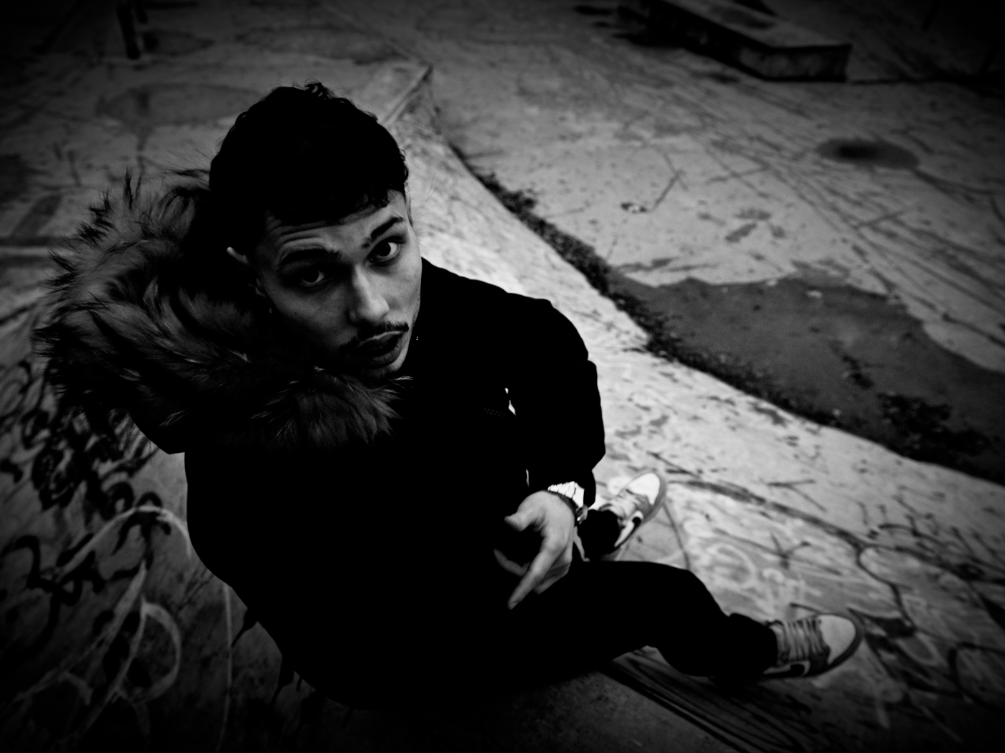 A woman with short dark hair, wearing a hoodie with a fur-lined hood, is sitting on a graffiti-covered surface, looking up at the camera with a serious expression in a black-and-white photo.