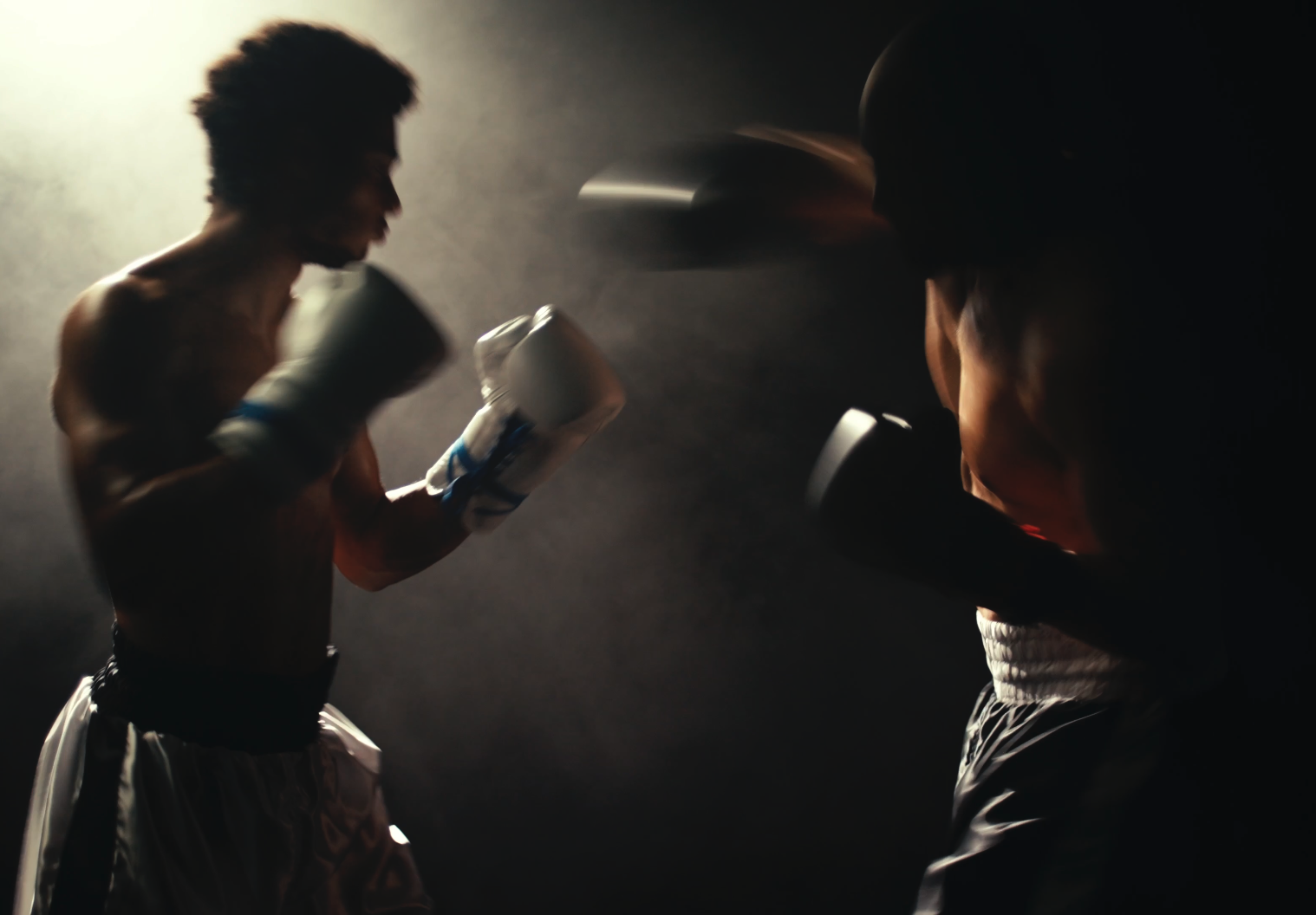 Two boxers training in a dark setting, facing each other with their fists raised, one in white gloves, both shirtless and wearing boxing shorts.