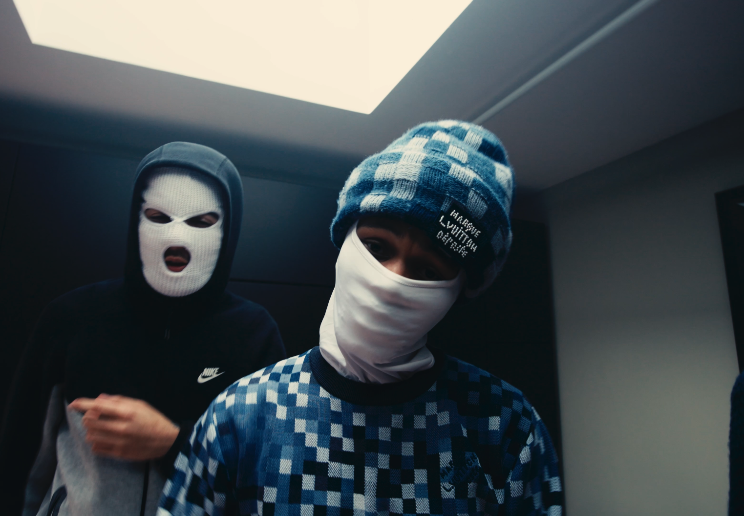 Two young men wearing masks and hoodies, one with a ski mask and the other with a face covering, indoors with a ceiling light overhead.