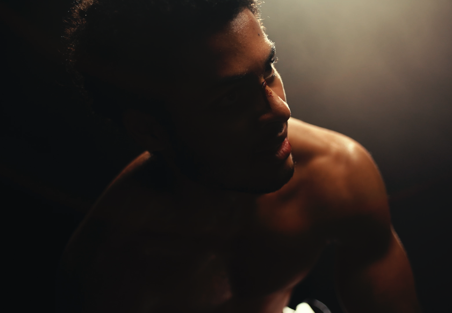 A shirtless man with dark skin and curly hair leaning forward in a dimly lit space, with light shining on part of his face and shoulder.
