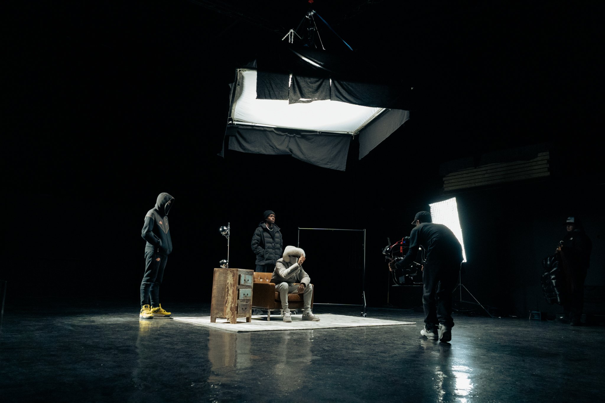 Film set with a seated person wearing a hoodie and headphones, surrounded by crew members and camera equipment, under large softbox lights.