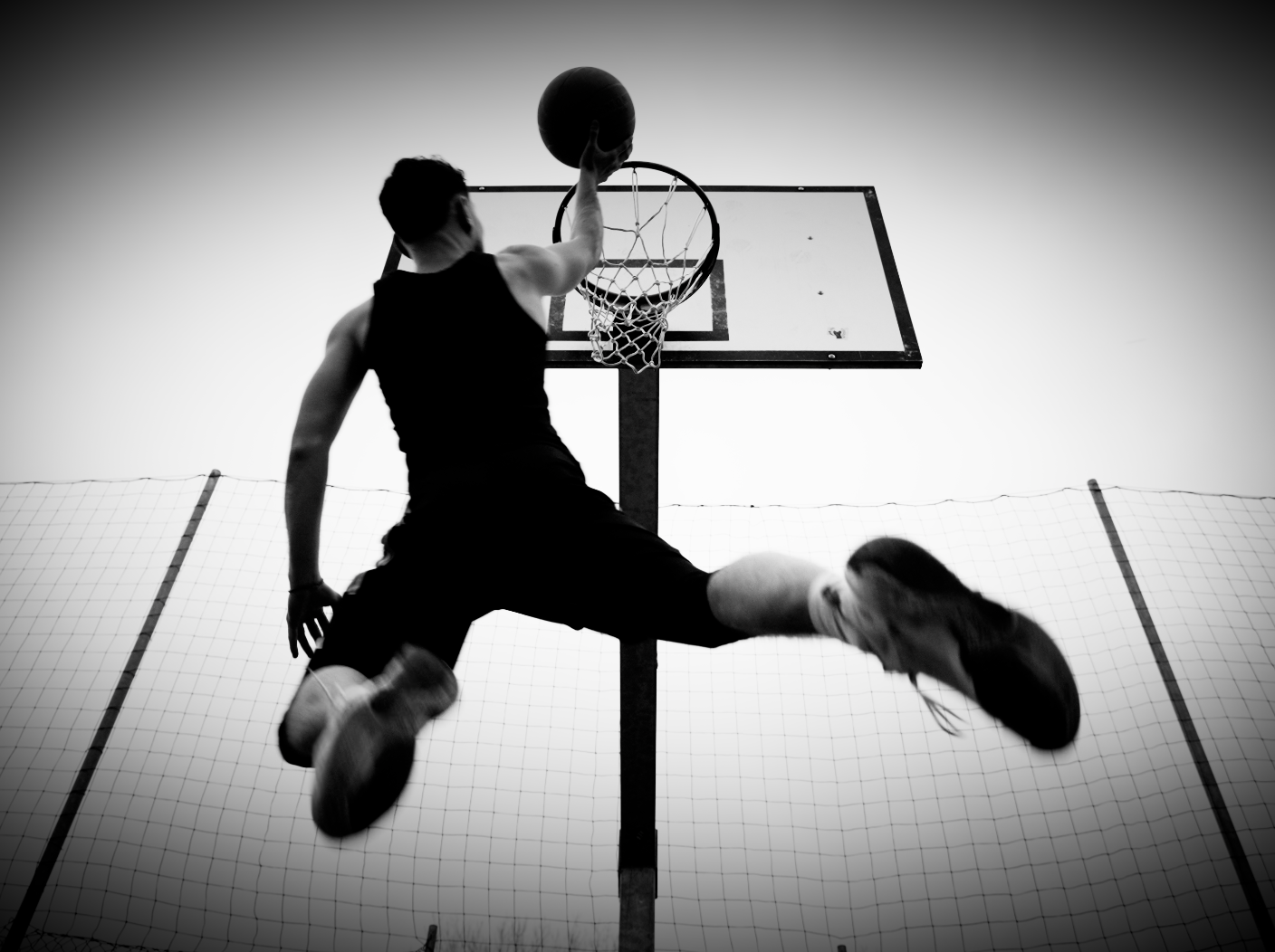 A person playing basketball, flying towards the hoop for a dunk. Black and white image, with the individual in a black tank top and shorts, captured mid-air with a basketball in hand.