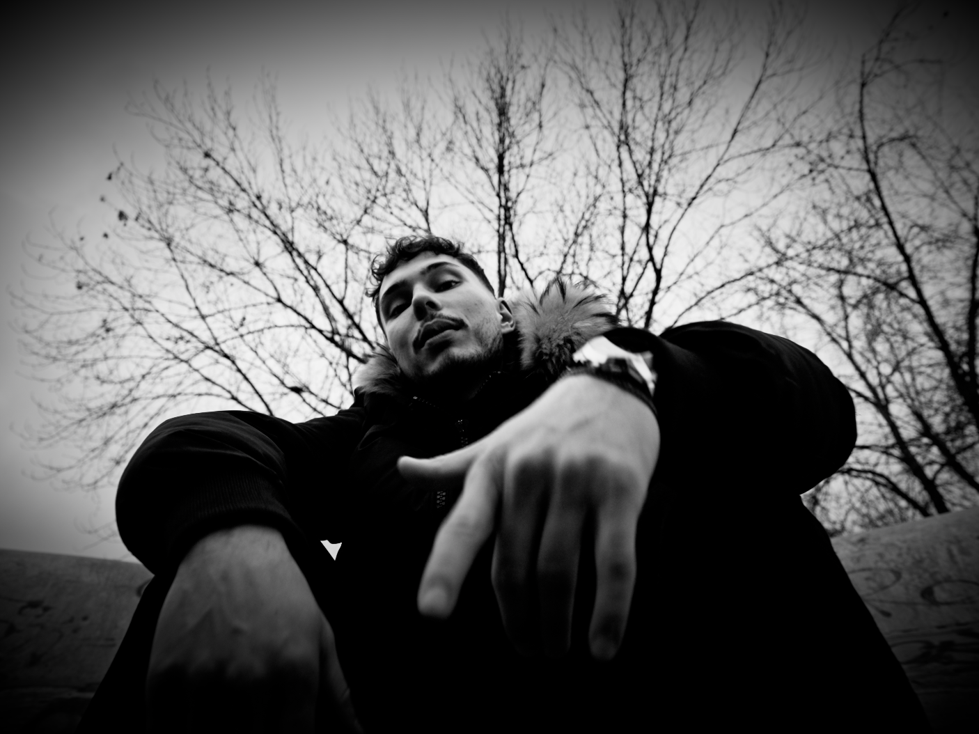 A young man in a dark jacket with a fur-lined hood leaning forward, pointing towards the camera, with leafless trees in the background, captured in black and white.