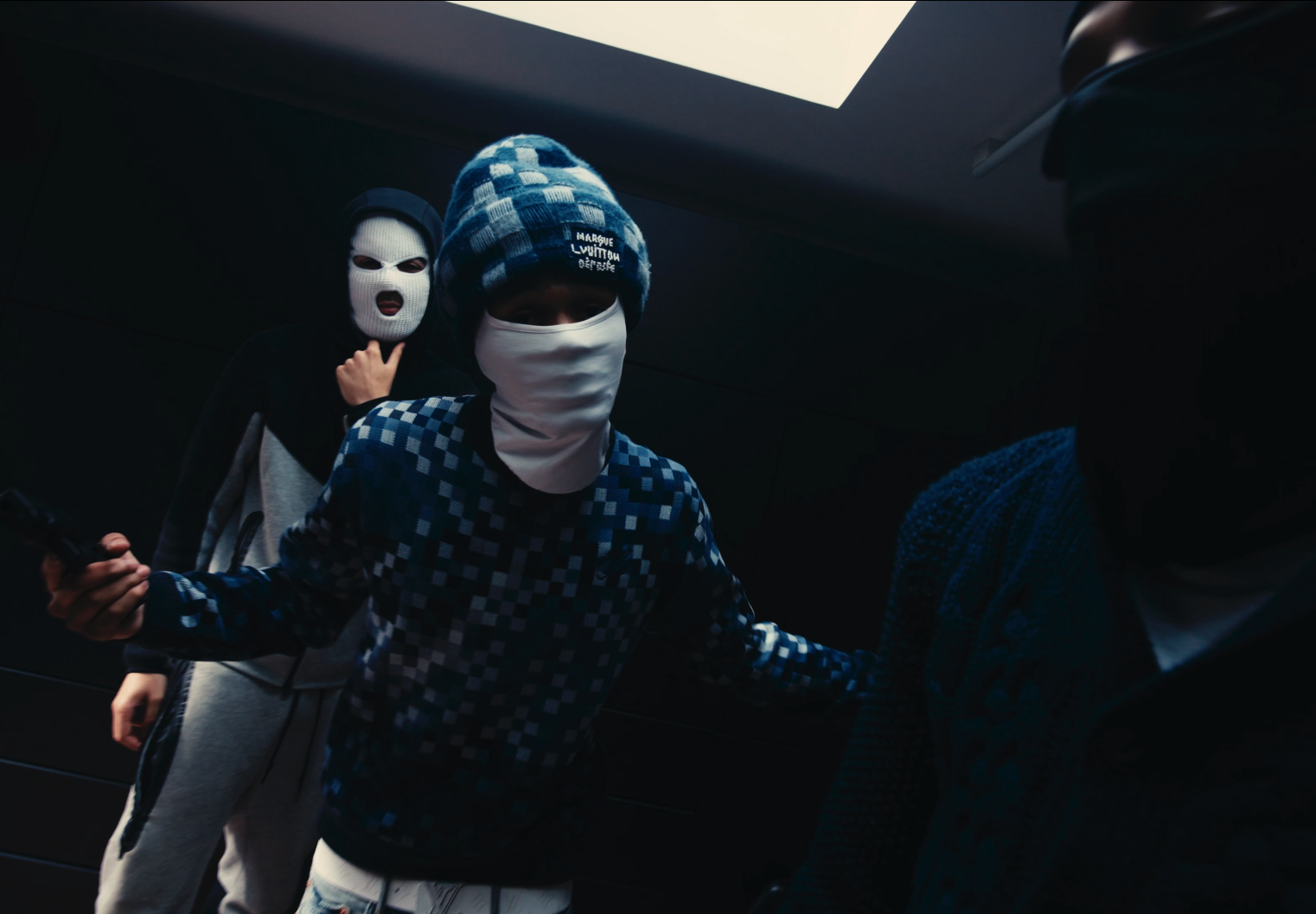 Three individuals wearing masks in a dark environment, one with a skull mask, one with a white face mask and a beanie, and one with a black face mask, engaged in a confrontational stance.