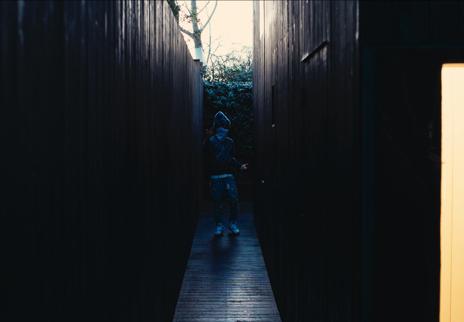 A person standing in a narrow outdoor wooden passageway between dark wooden walls, illuminated by natural light at the end, with trees and bushes in the background, wearing a hoodie, face mask, jeans, and sneakers.