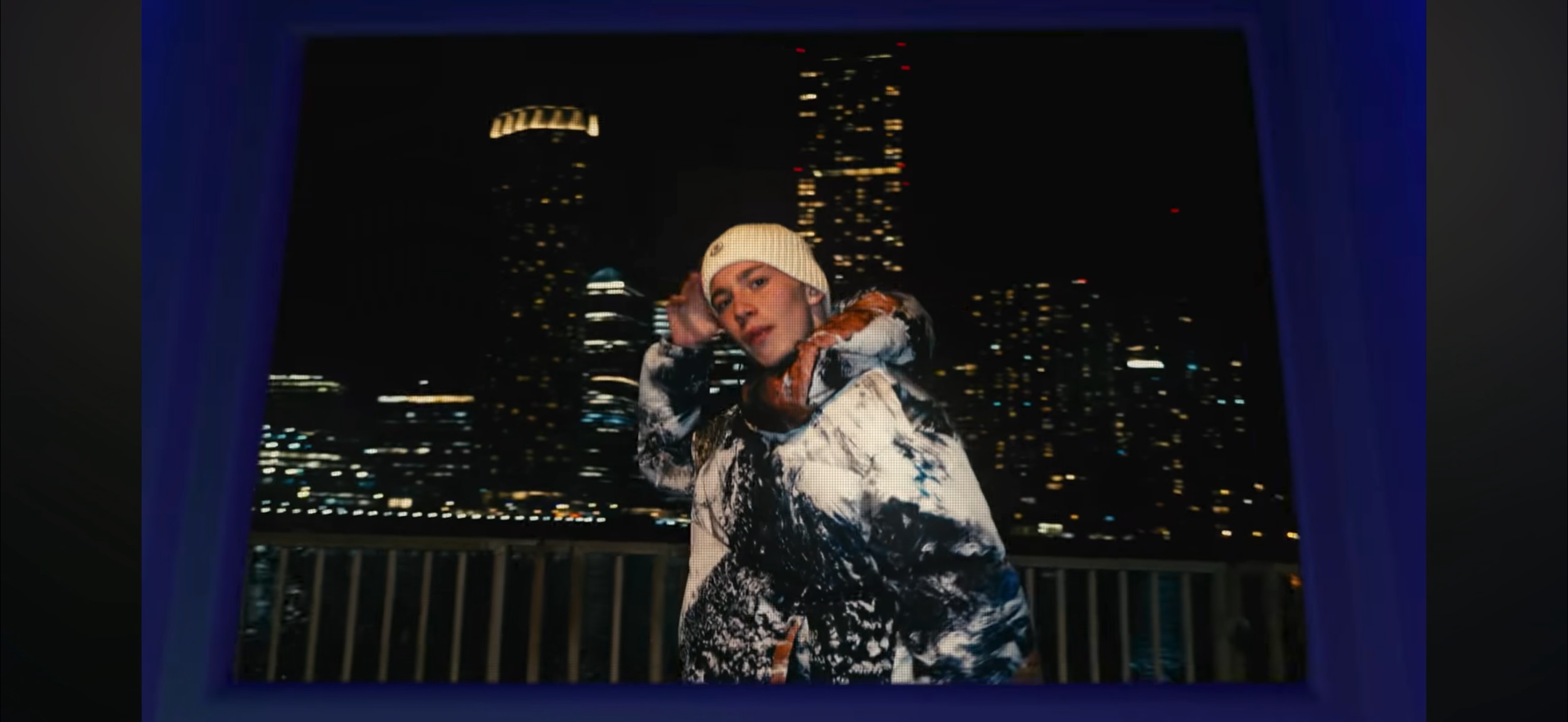 A young person standing on a balcony at night, with a city skyline and tall buildings with lights in the background. They are wearing a white beanie and a patterned jacket.