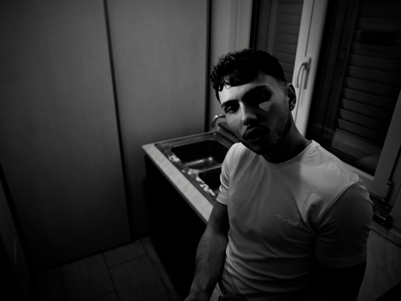 A young man with short curly hair and earrings standing in a kitchen with a sink and cabinets, looking at the camera in black and white.