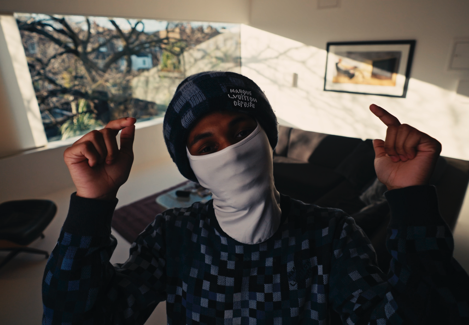 A person wearing a black and blue checkered jacket, a black and blue striped beanie, and a white face mask, standing indoors with hands raised and fingers pointing upward. Behind them is a large window showing trees outside, and a sofa with pillows i