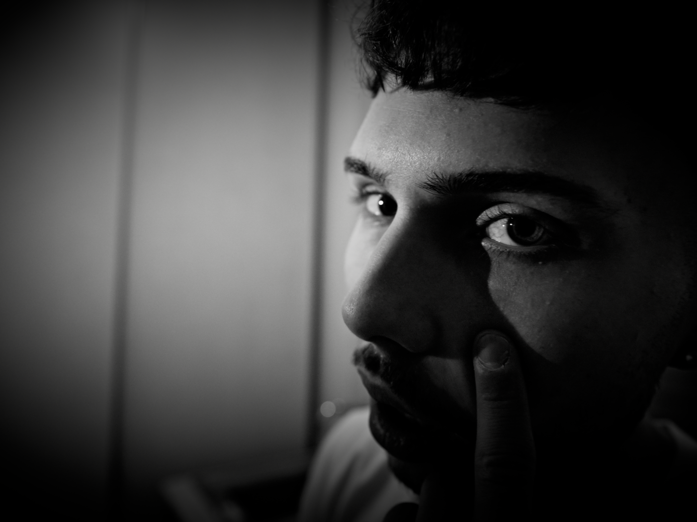 Close-up black and white photo of a young man with intense gaze, touching his face with his finger, shadowed lighting highlights facial features.