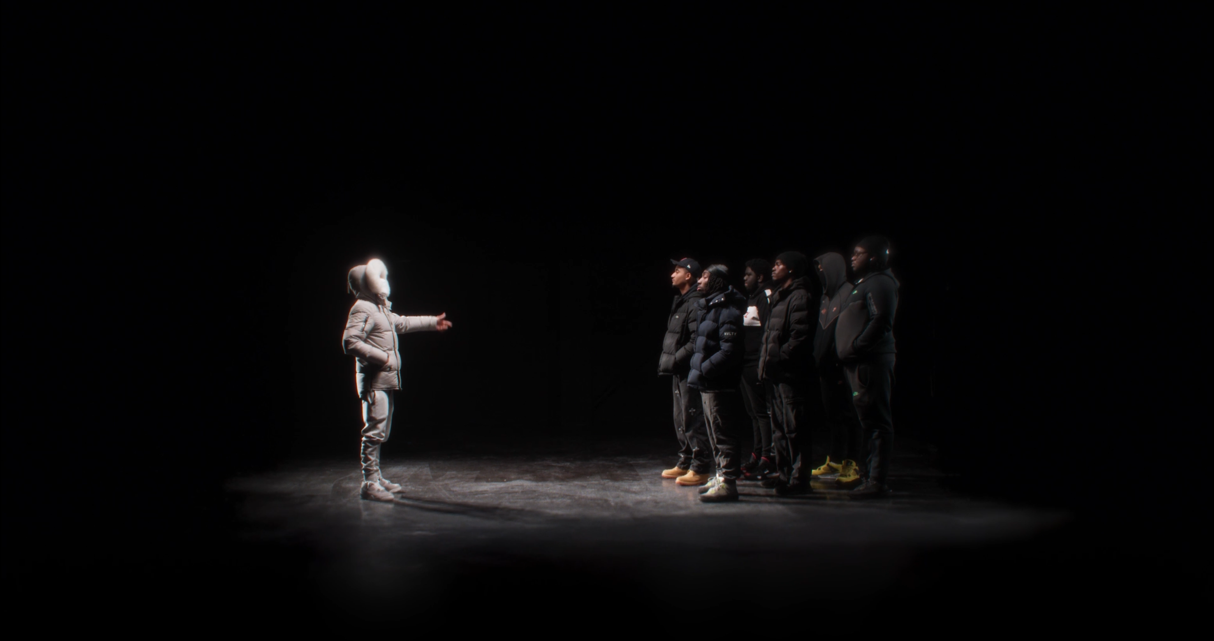 A woman with a hood and light-colored clothing speaks to a group of six people, all dressed warmly in dark jackets, on a dark stage with a spotlight highlighting them.