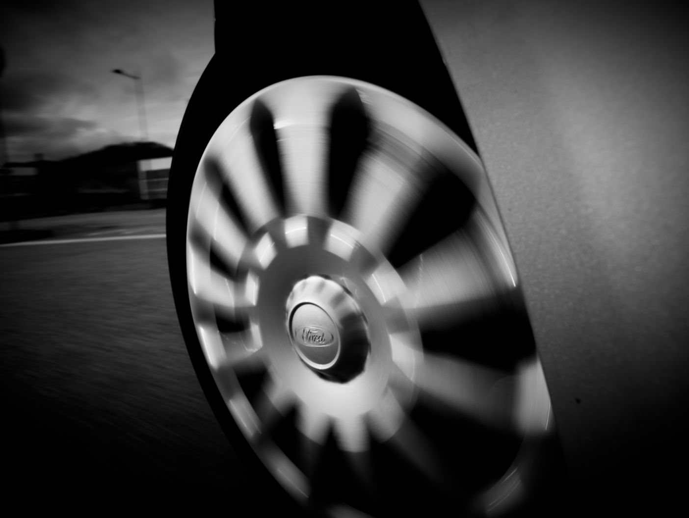 Close-up of a car wheel in motion, with the Ford logo visible on the hubcap, captured in black and white.