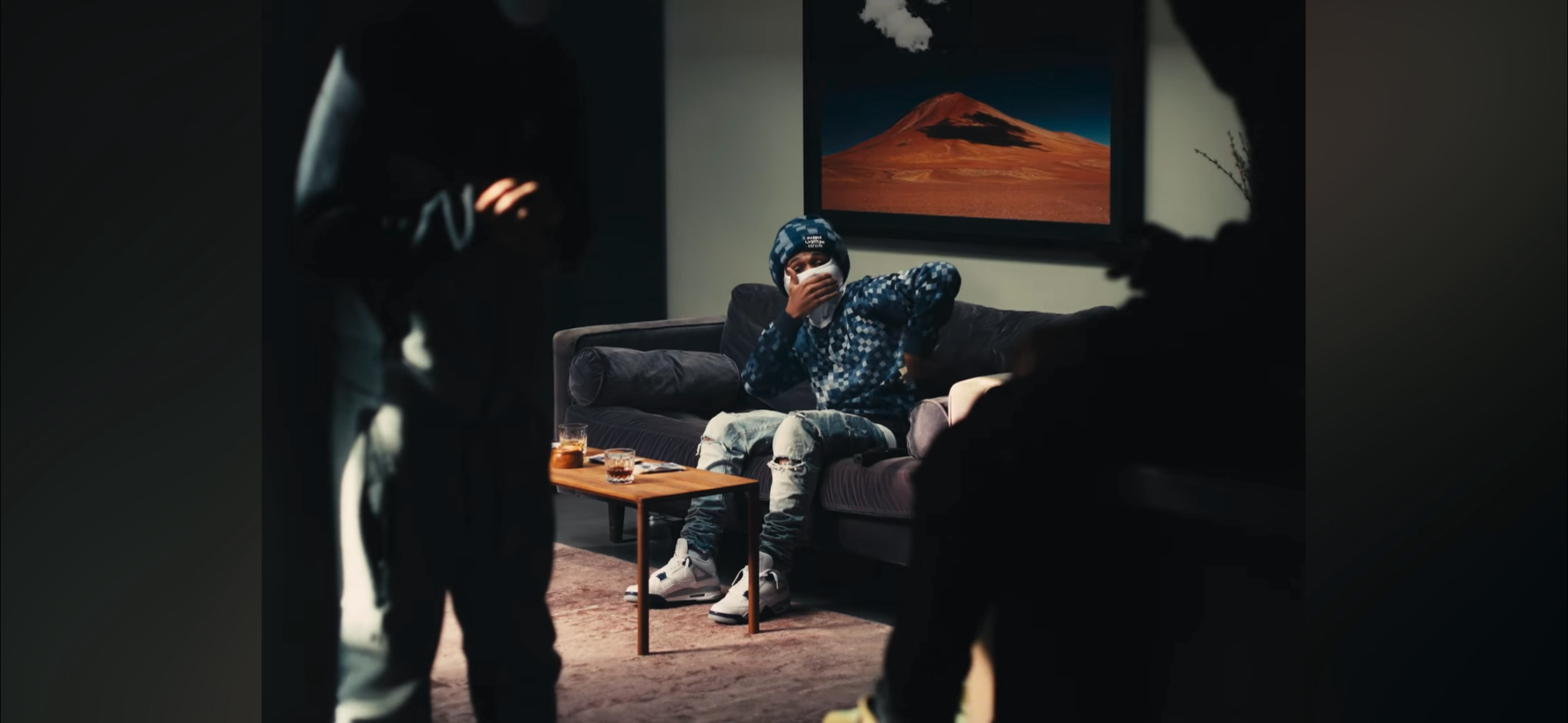 A boy sitting on a gray couch in a dark living room, wearing a blue patterned hoodie, ripped jeans, white sneakers, and a matching beanie, covering his mouth with his hand. There are two glasses of drinks on a wooden coffee table in front of him. Two other people, partially visible and out of focus, stand nearby. A framed picture of a mountain hangs on the wall behind him.