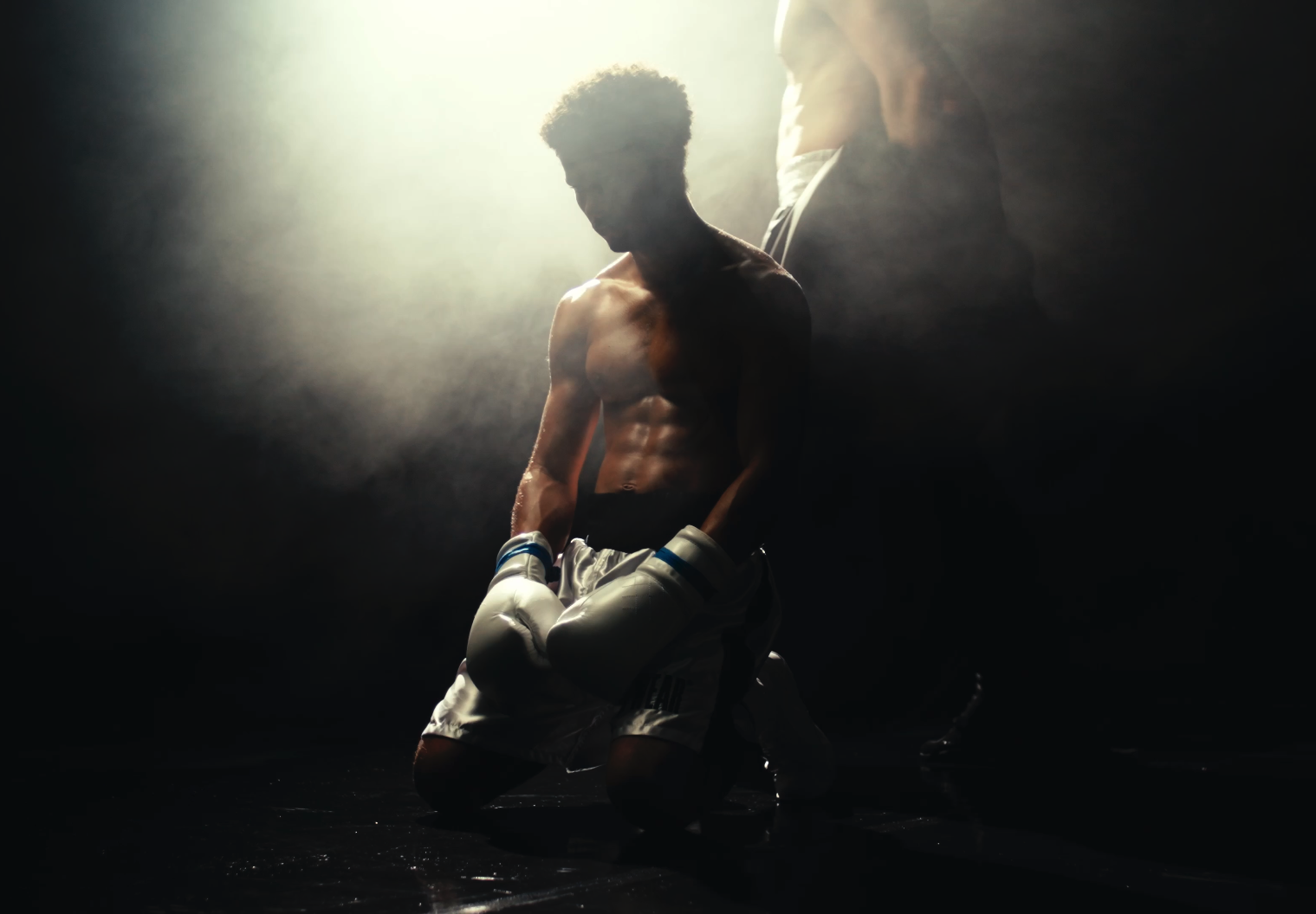 A shirtless male boxer wearing boxing gloves and shorts kneeling on the ground in a dimly lit environment, with a bright light source behind him creating a silhouette.