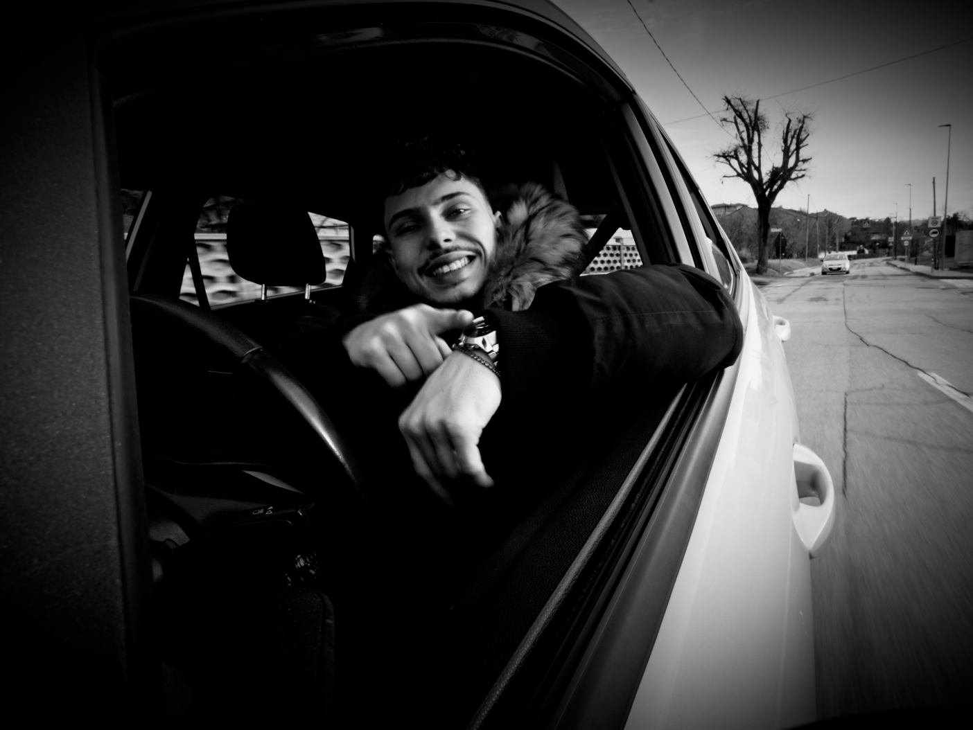 Black-and-white photo of a man leaning out of a car window, smiling and pointing at his watch, with a tree and street in the background.