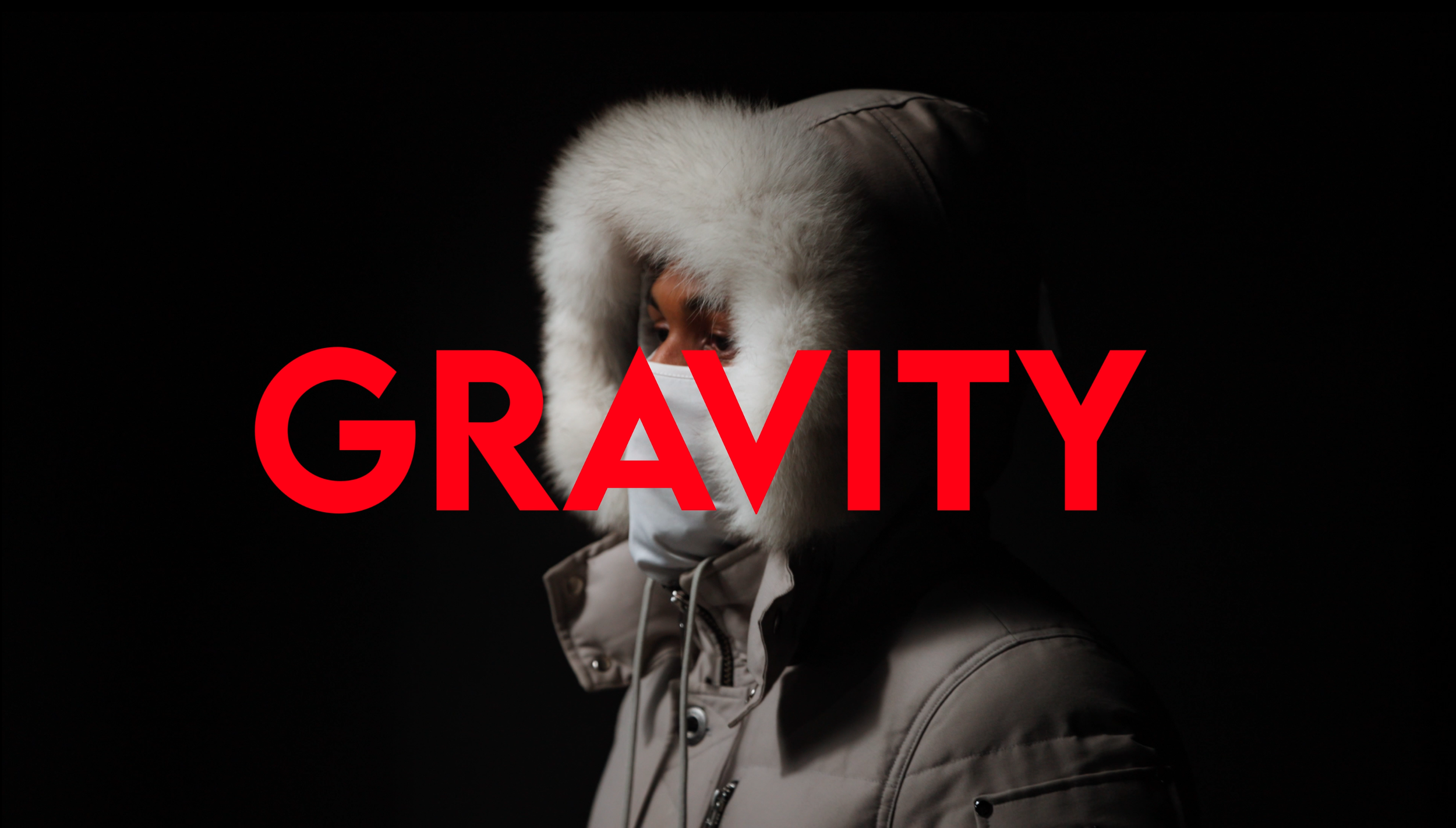Person wearing a winter coat with a fur-lined hood, a face mask, and looking to the side, against a dark background. The word 'GRAVITY' is written in large red letters across the image.