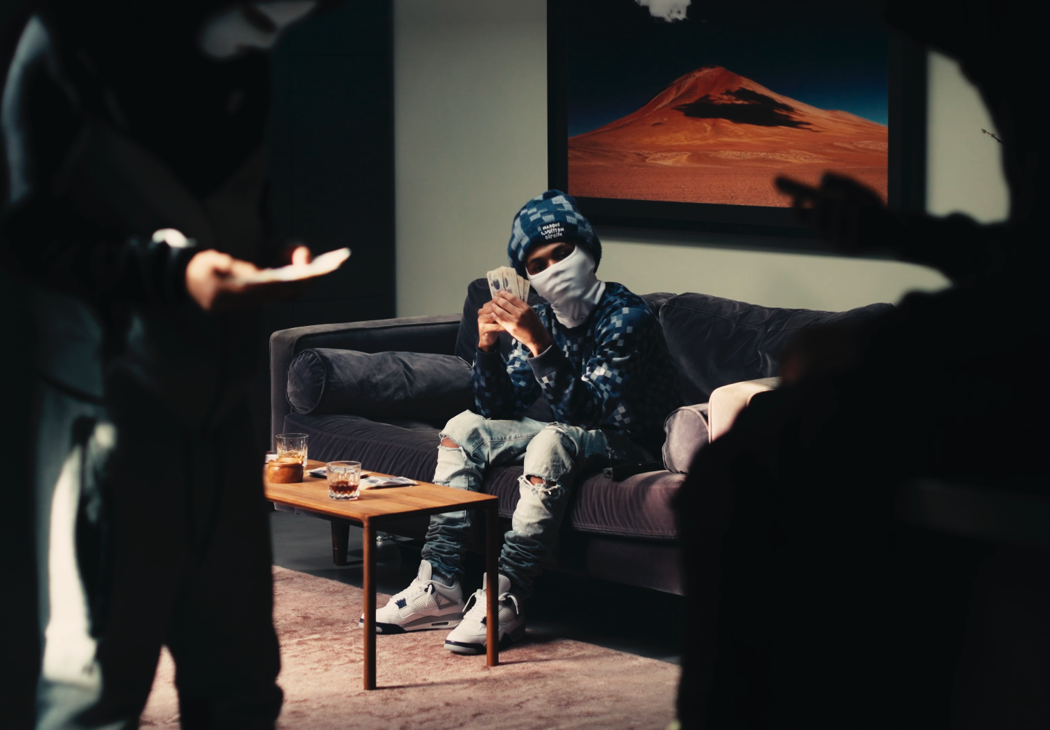 A young man wearing a mask, blue hoodie, and ripped jeans sitting on a sofa, holding cash. Two masked individuals are in the foreground, one with a phone. There are drinks on a small wooden table and a mountain painting on the wall.