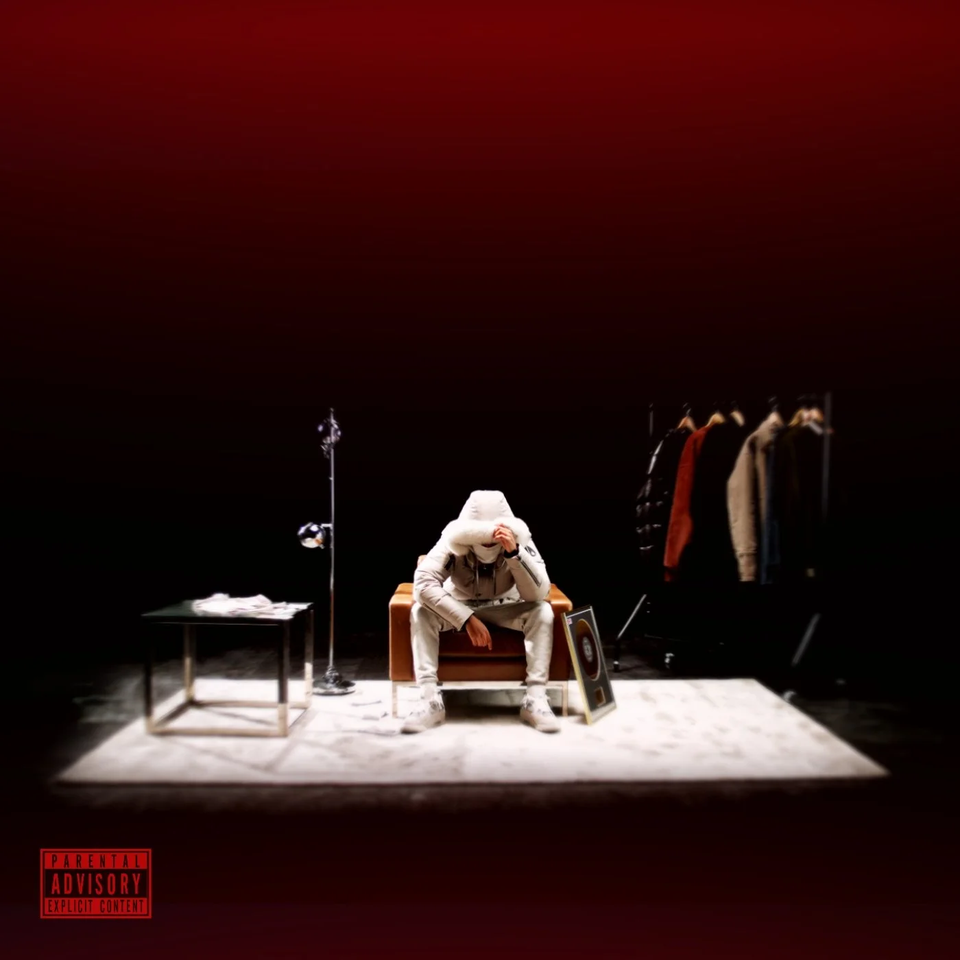 A person sitting on a brown sofa with their hands covering their face, dressed in light-colored streetwear and a hoodie. They are in a room with a dark background and minimal furniture, including a small black table with papers, a coat rack with jackets, a framed picture on the floor, and some studio lighting equipment. A parental advisory label is in the bottom left corner.