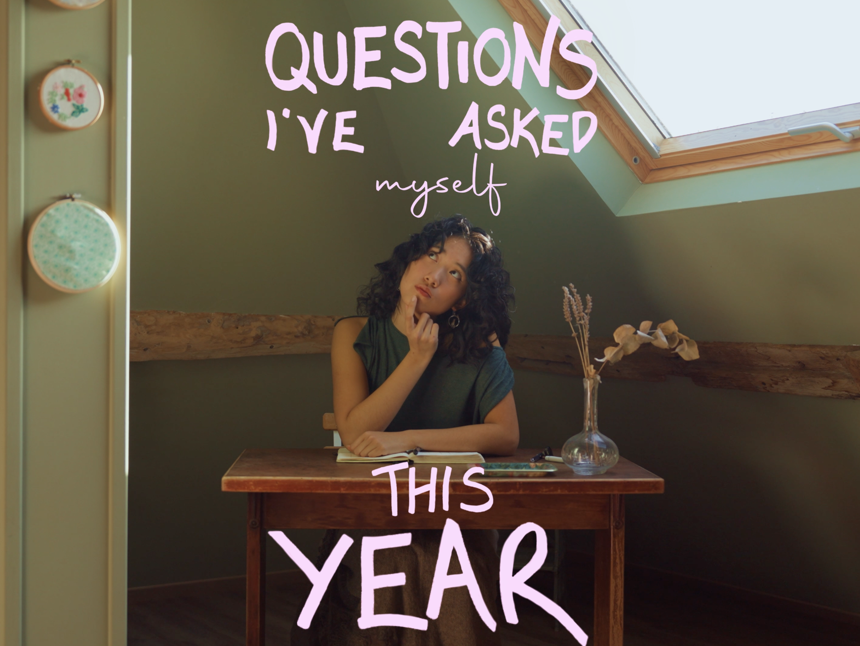 A woman sitting at a wooden desk with a notebook, pen, and vase of dried flowers, looking up thoughtfully next to a window in an attic room with green walls, with text overlay that reads, "Questions I've Asked Myself This Year."