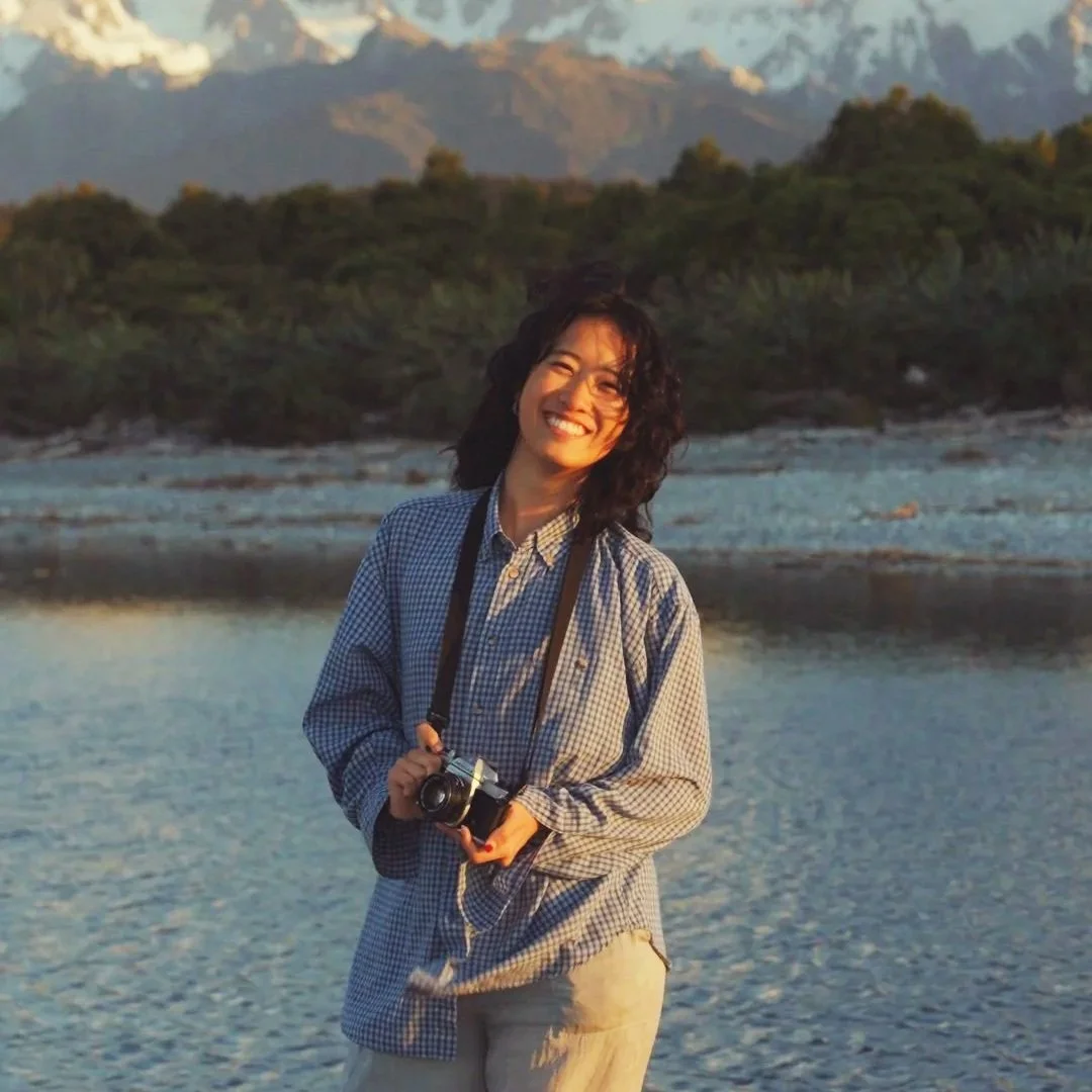 A woman with curly dark hair smiling at the camera, holding a camera in her hand, standing outdoors by a body of water with mountains and trees in the background during sunset.