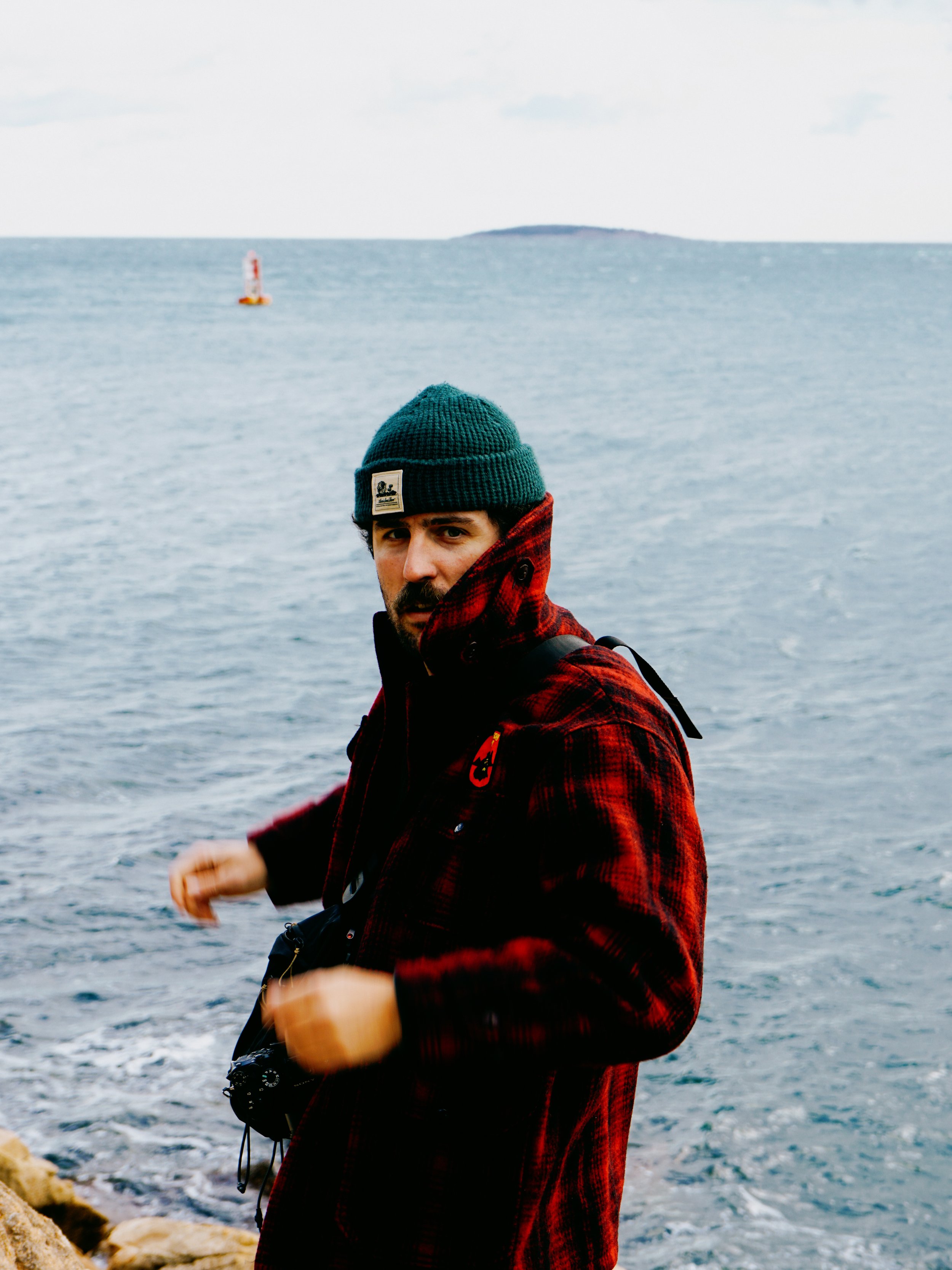 A man with dark hair and a beard dressed in a red and black plaid jacket, green beanie, and red scarf, standing near water with a small island and a buoy in the background.