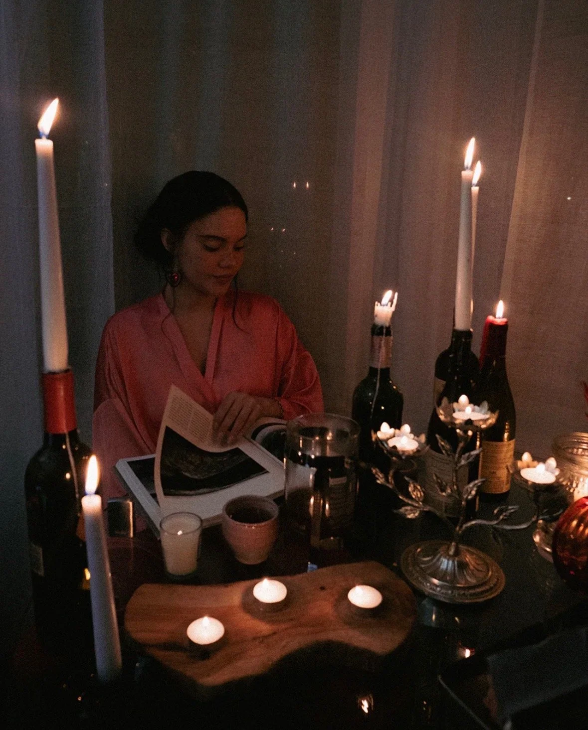 A woman in a pink satin robe sitting at a dinner table lit by candles, reading a magazine or book in a dimly lit room.