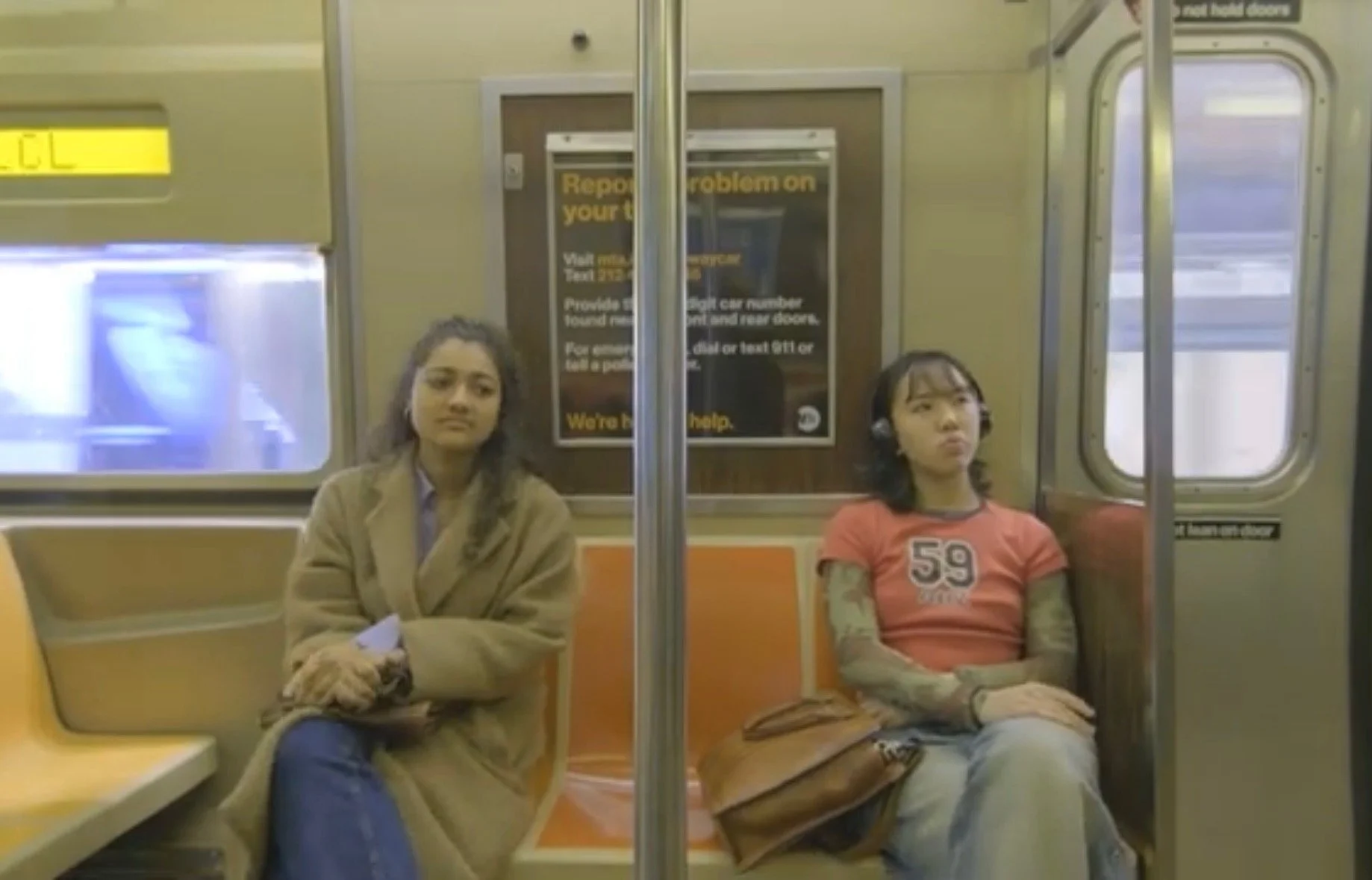 Two young women sitting apart on subway seats, one in an orange T-shirt with tattooed arms and headphones, the other in a tan coat with crossed arms.