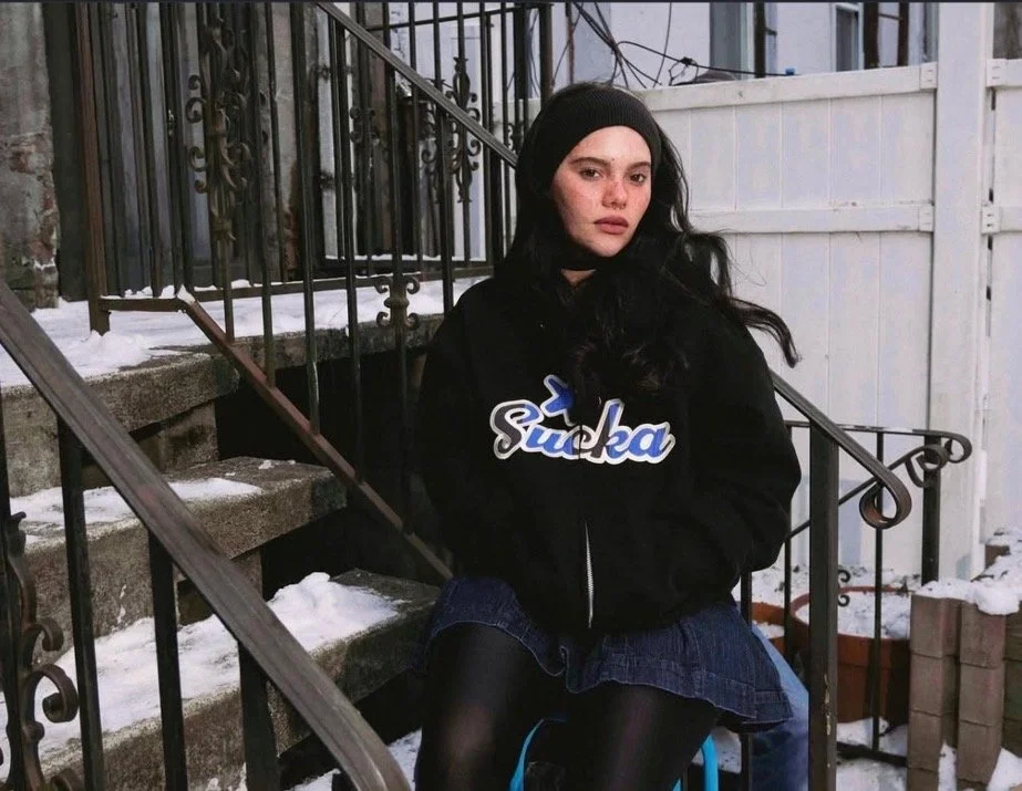 A young woman with black hair and a black headband sitting on outdoor stairs in winter, wearing a black hoodie with the word 'Suka' in blue and white, a denim skirt, and black tights, in a residential area.